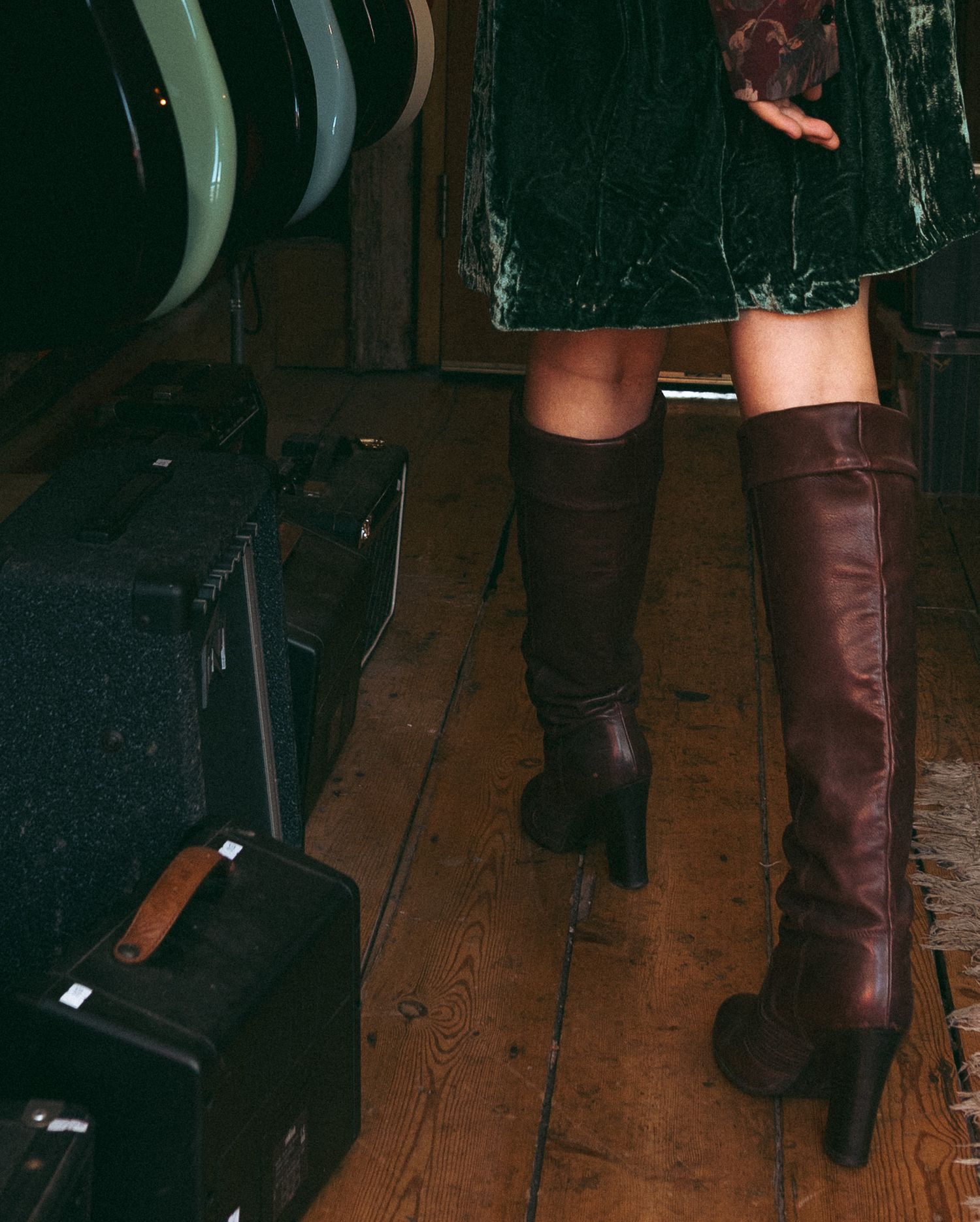 Woman wearing dark brown knee-high leather heeled boots with a green velvet skirt on rustic wooden floors.