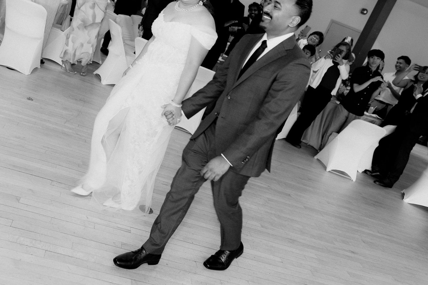 Black and white photo of a smiling groom in a suit dancing with a bride in a white gown while wedding guests watch from tables.