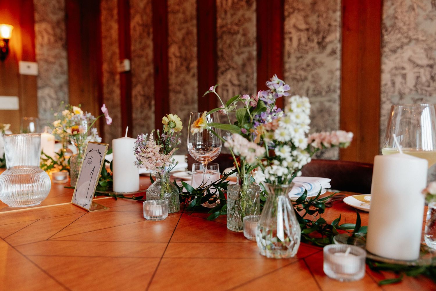 Elegant wedding reception table decorated with white candles, glass vases, wildflowers and greenery on a polished terracotta tile surface.