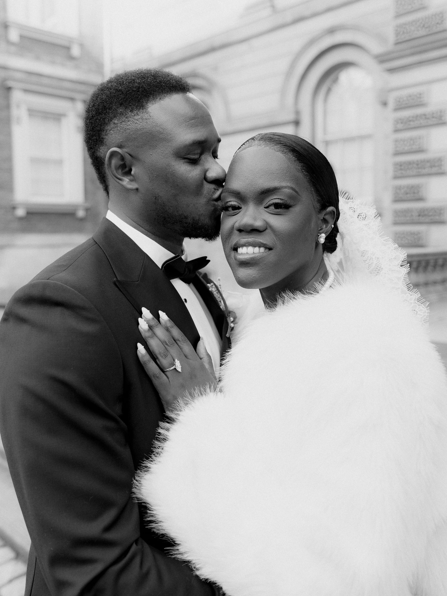 Black and white wedding portrait of a groom in a tuxedo kissing his smiling bride in a fluffy white fur stole outside a historic building.