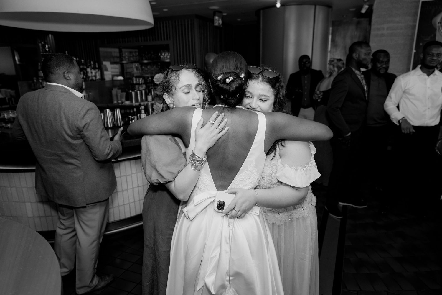 Bride in a white gown shares a warm embrace with two women at a wedding reception while guests mingle near the bar.