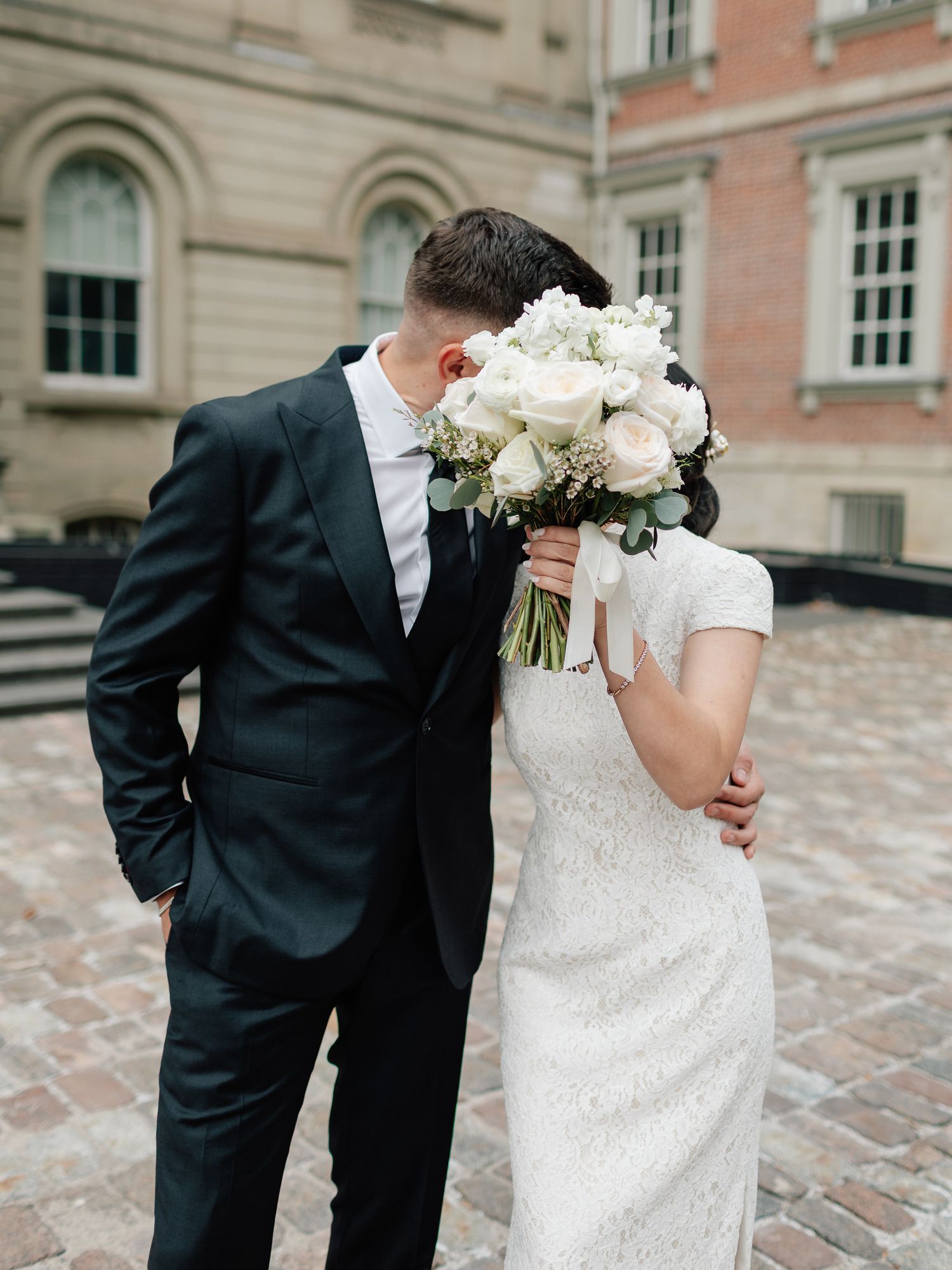 Bride and groom hide their kiss behind a white rose bouquet on a cobblestone courtyard, he in a dark suit and she in a lace dress.
