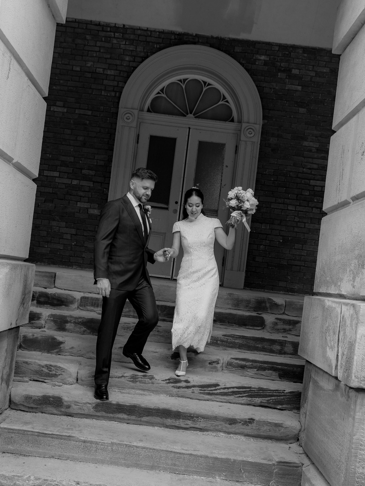 Black and white photo of a bride and groom holding hands and walking down stone steps outside a brick building with an arched doorway.