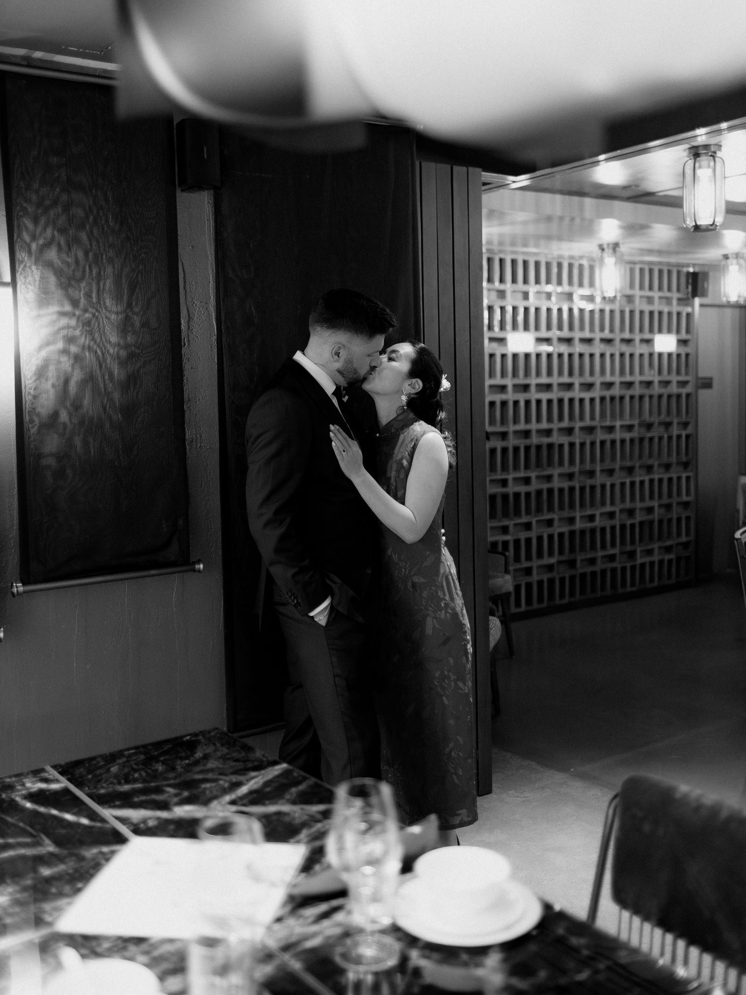 Black and white photo of a couple kissing in an elegant restaurant beside a marble table set with glasses and plates.