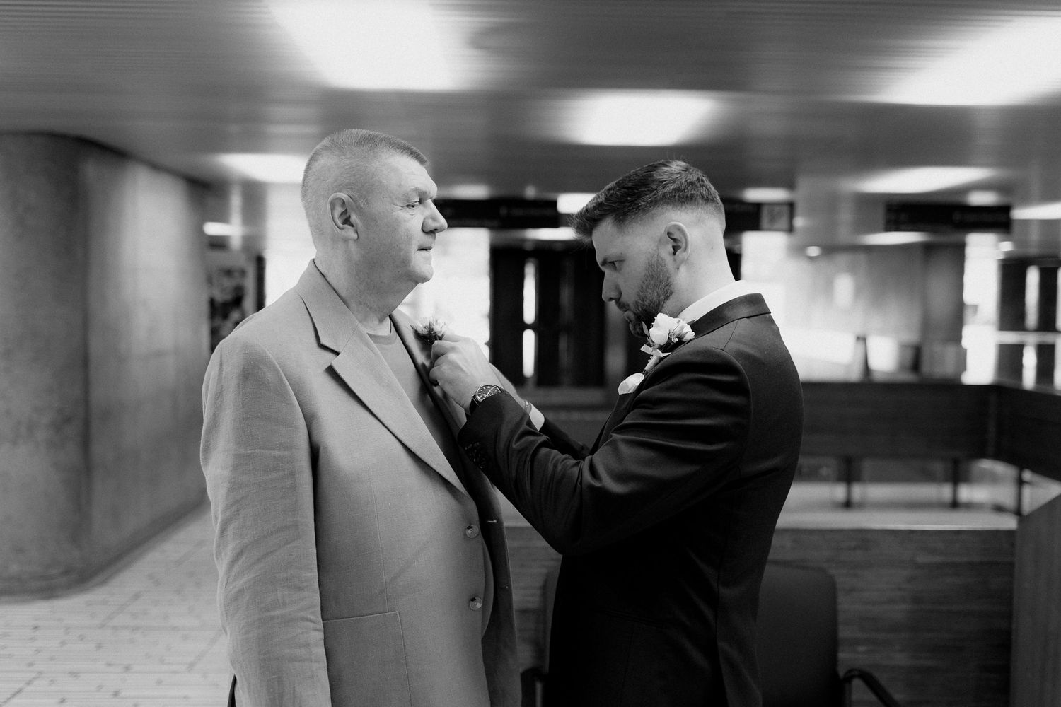 Groom in a dark suit pins a boutonniere onto a man's light jacket in a black and white wedding photo taken indoors.