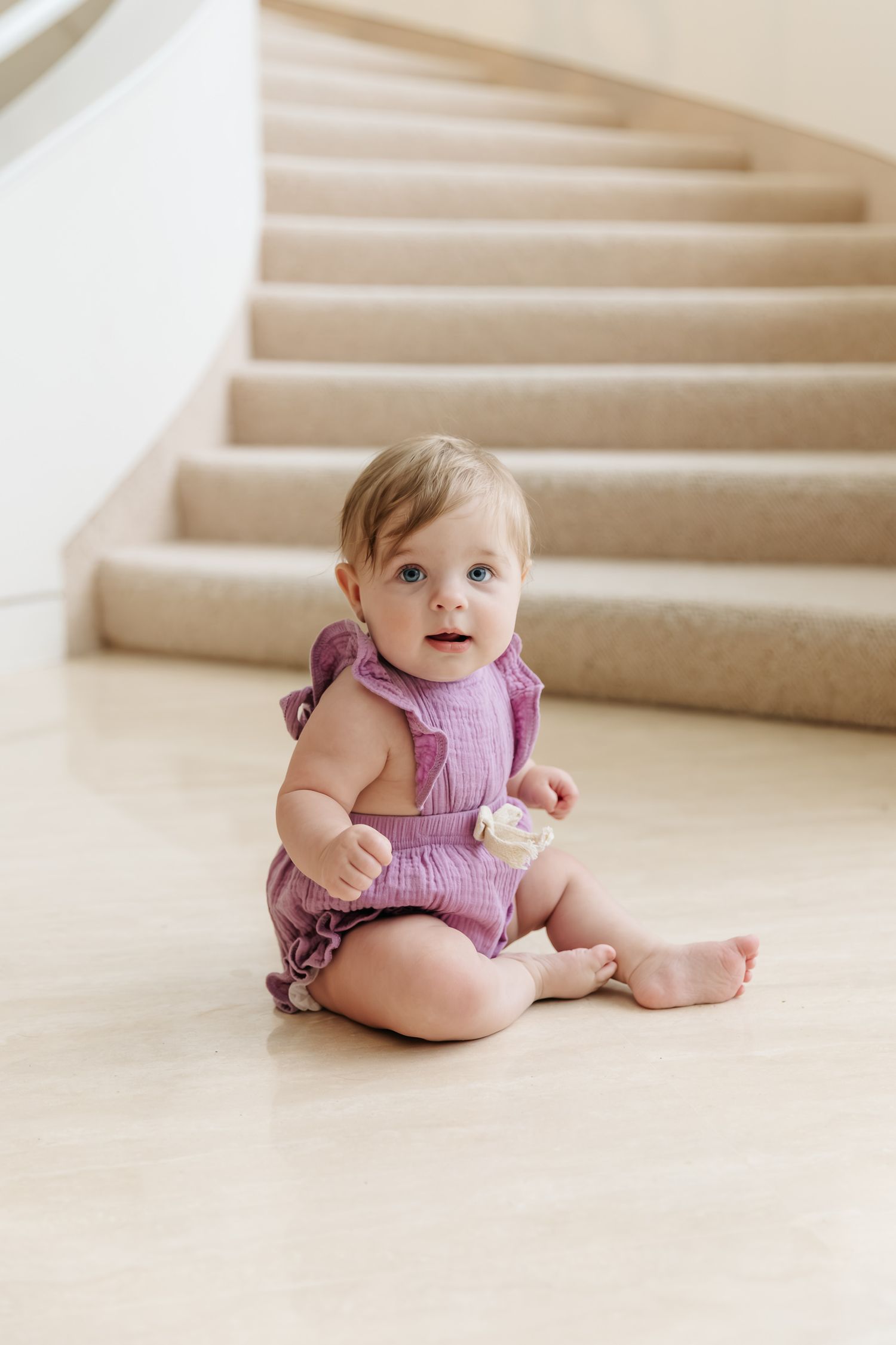 Baby in a purple ruffled romper sits on a smooth marble floor holding a small toy, with a carpeted curved staircase behind.