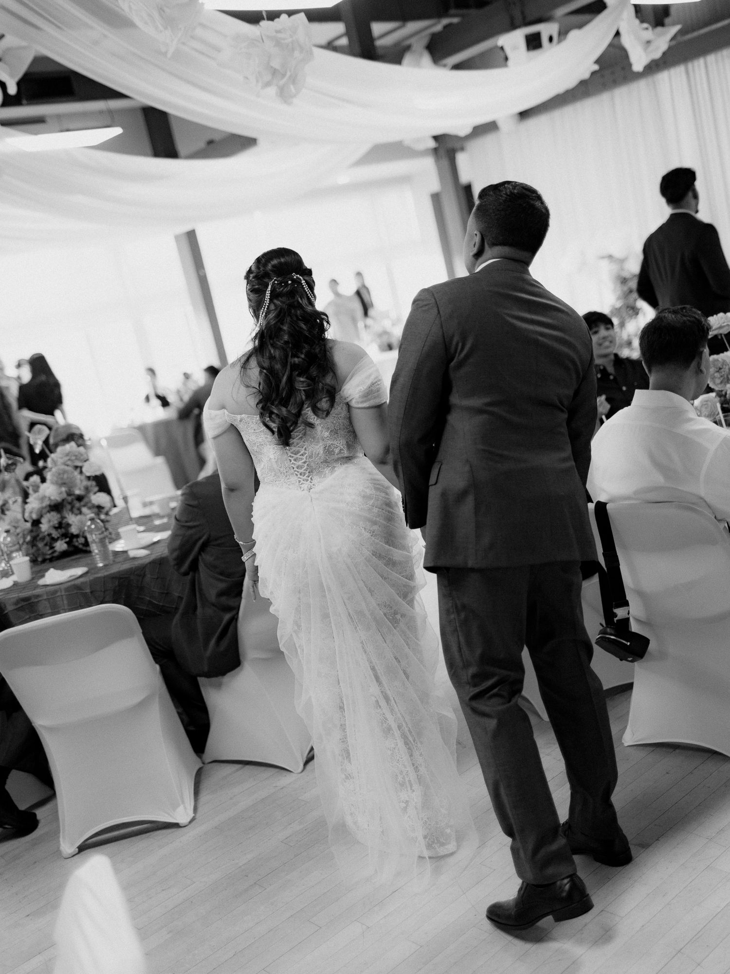 Black and white photo of a bride in a lace wedding gown walking beside a groom in a suit through a reception hall with draped fabric.
