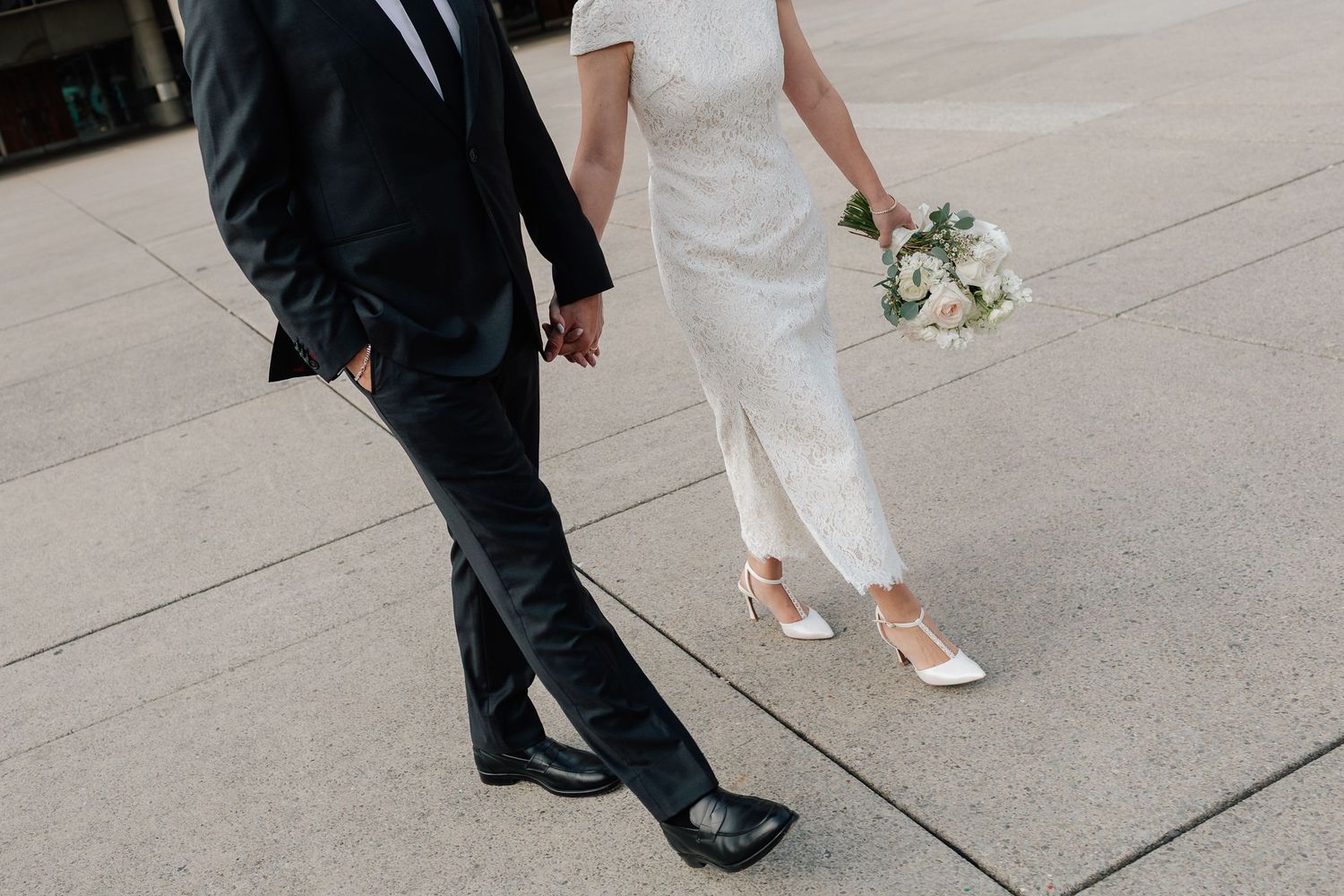 Bride and groom walking hand in hand, bride holding white bouquet, wearing elegant wedding attire and white heels.