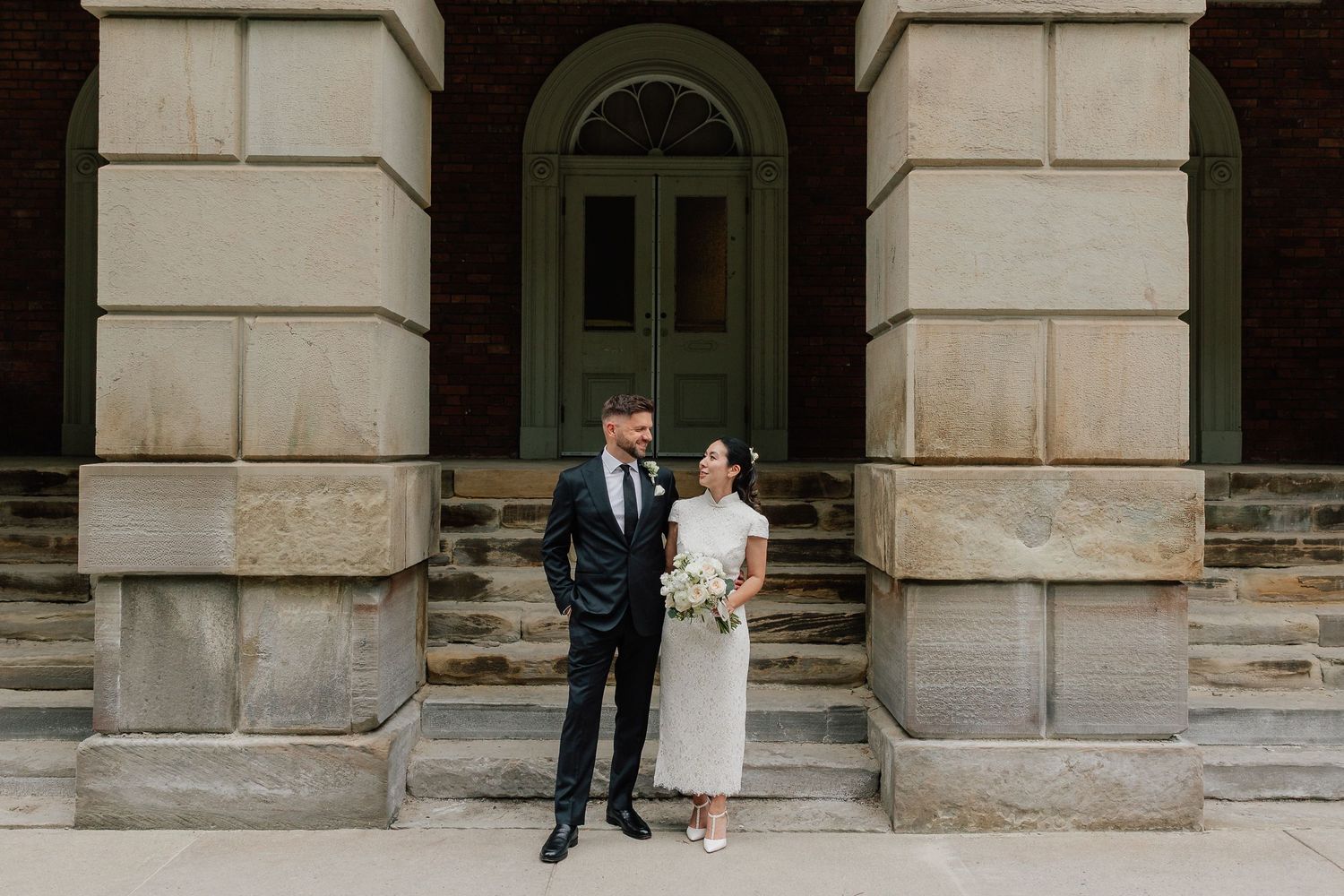 Bride in a white dress holding a bouquet stands beside a groom in a dark suit on stone steps in front of a historic brick building.