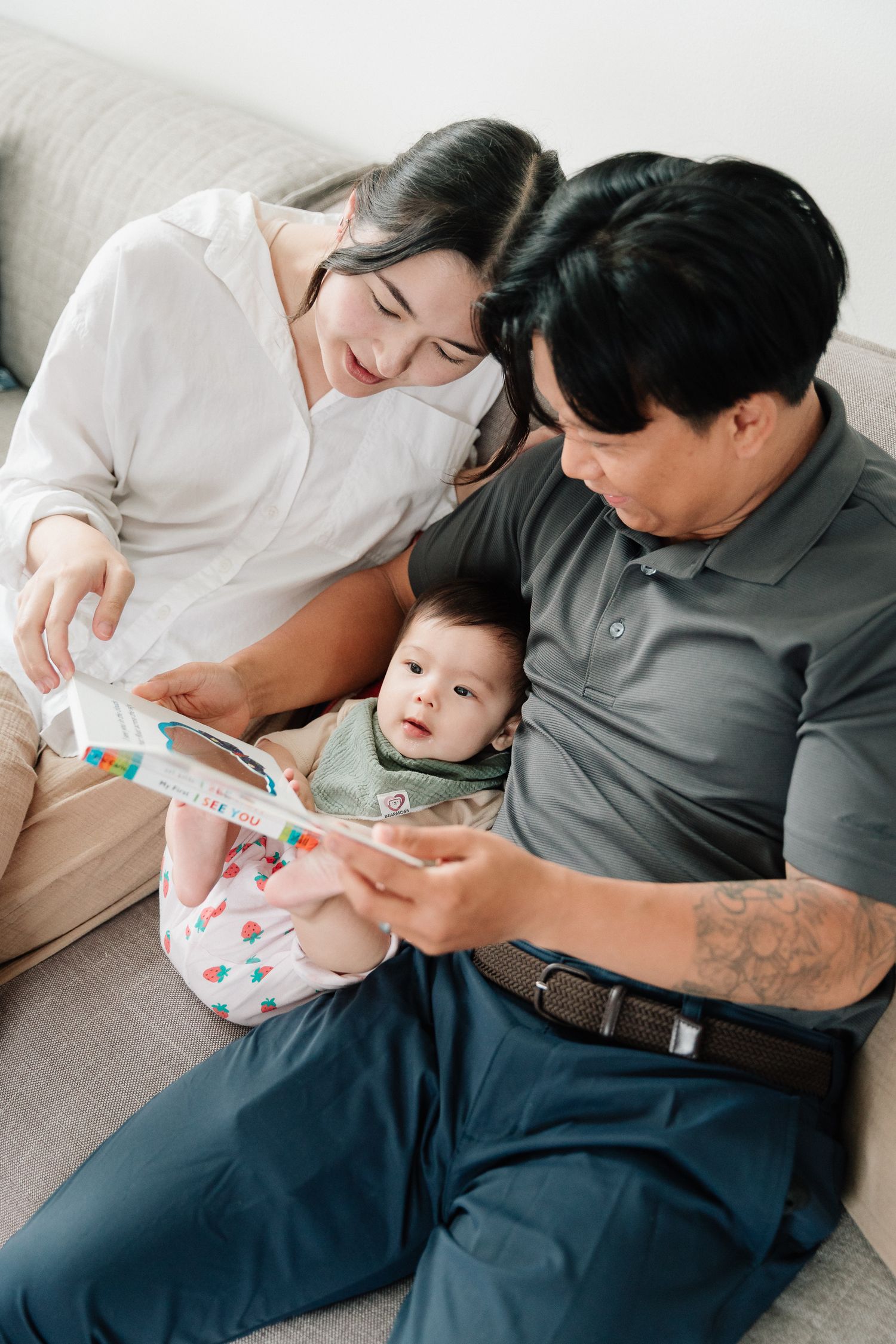 Smiling parents sit on a couch reading a colorful picture book with their baby, sharing a warm family bonding moment at home.