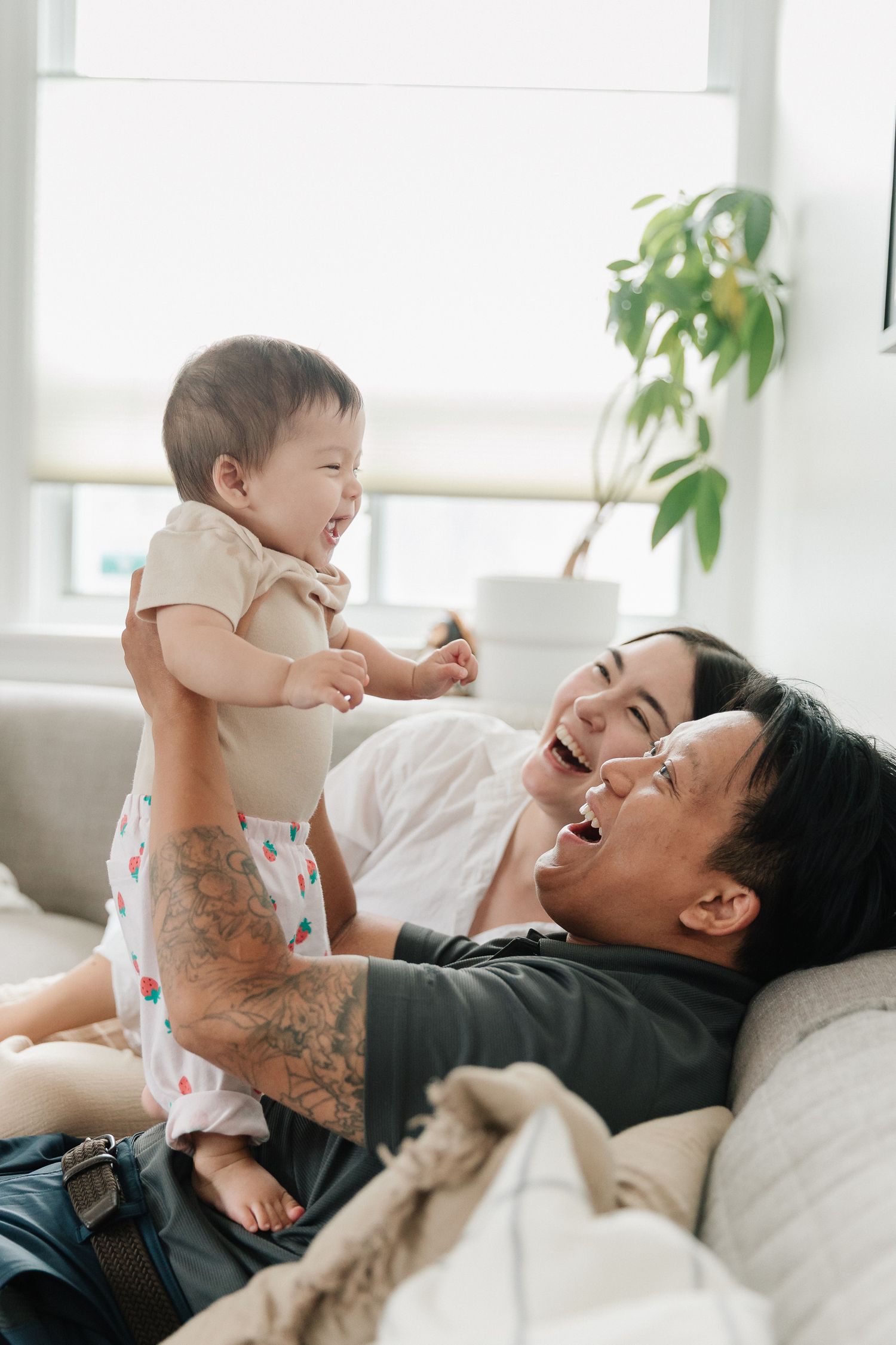 Smiling parents laugh on a sofa while a tattooed father lifts a giggling baby in the air in a bright, plant-filled living room.