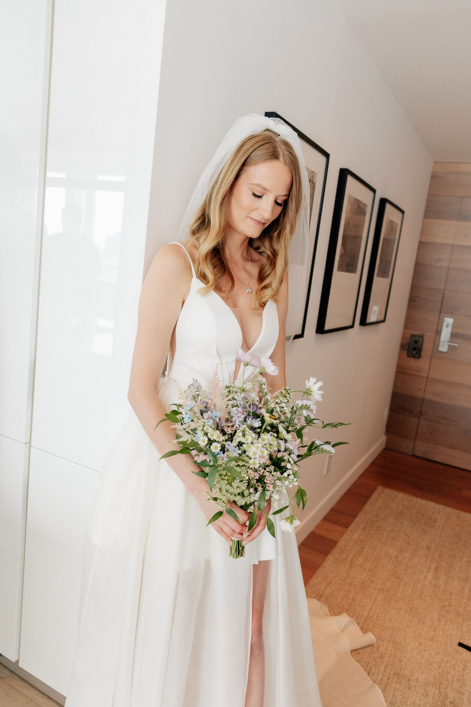Bride in a white gown and veil looks down at her wildflower bouquet while standing in a bright hallway with framed art.
