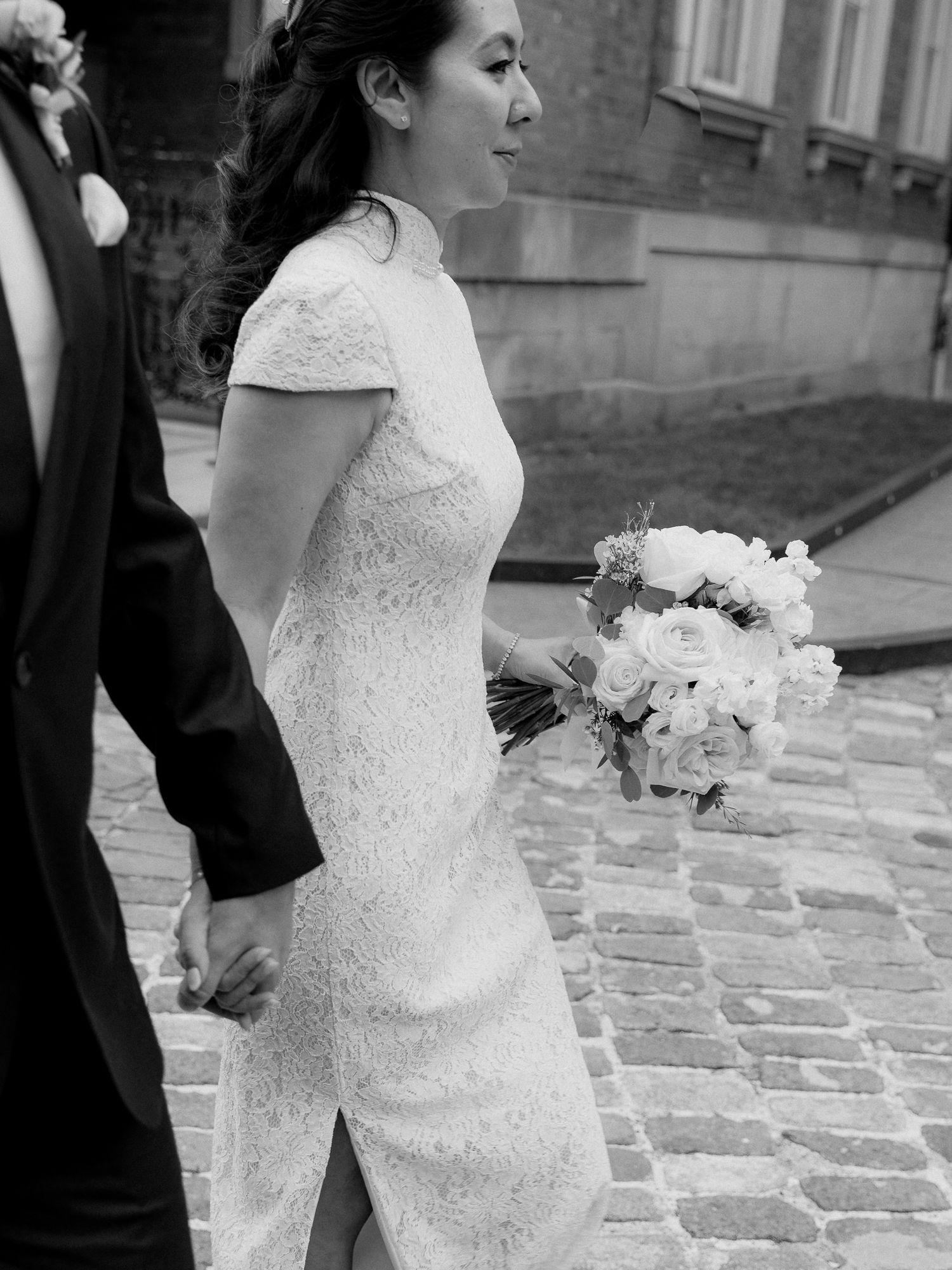 Bride in a lace qipao-style wedding dress holds a white rose bouquet and walks hand in hand with her groom on a cobblestone street.