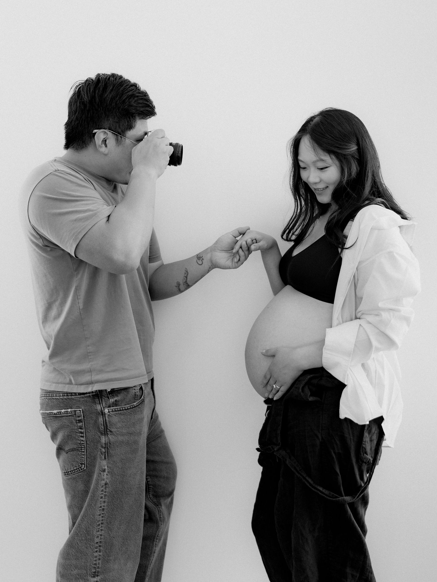 Black and white photo of a man photographing his smiling pregnant partner against a white background.