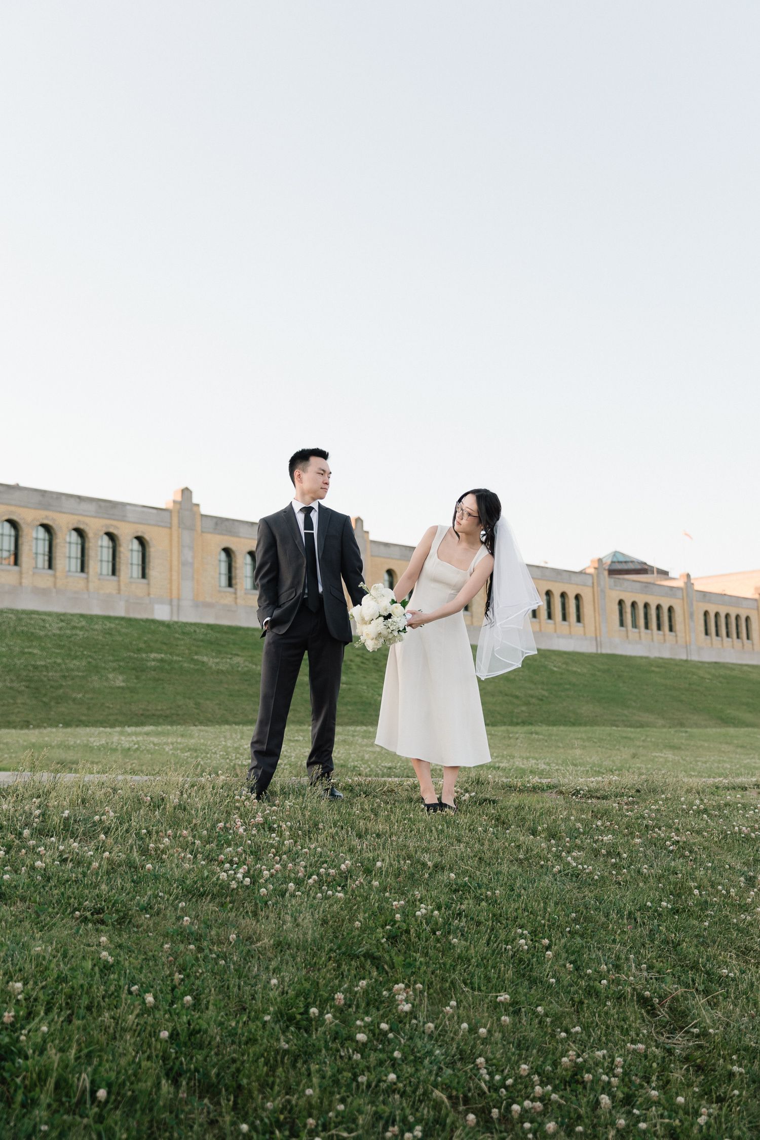 Bride in a white dress and veil holding a bouquet stands beside a groom in a dark suit on a grassy lawn in front of a historic building.