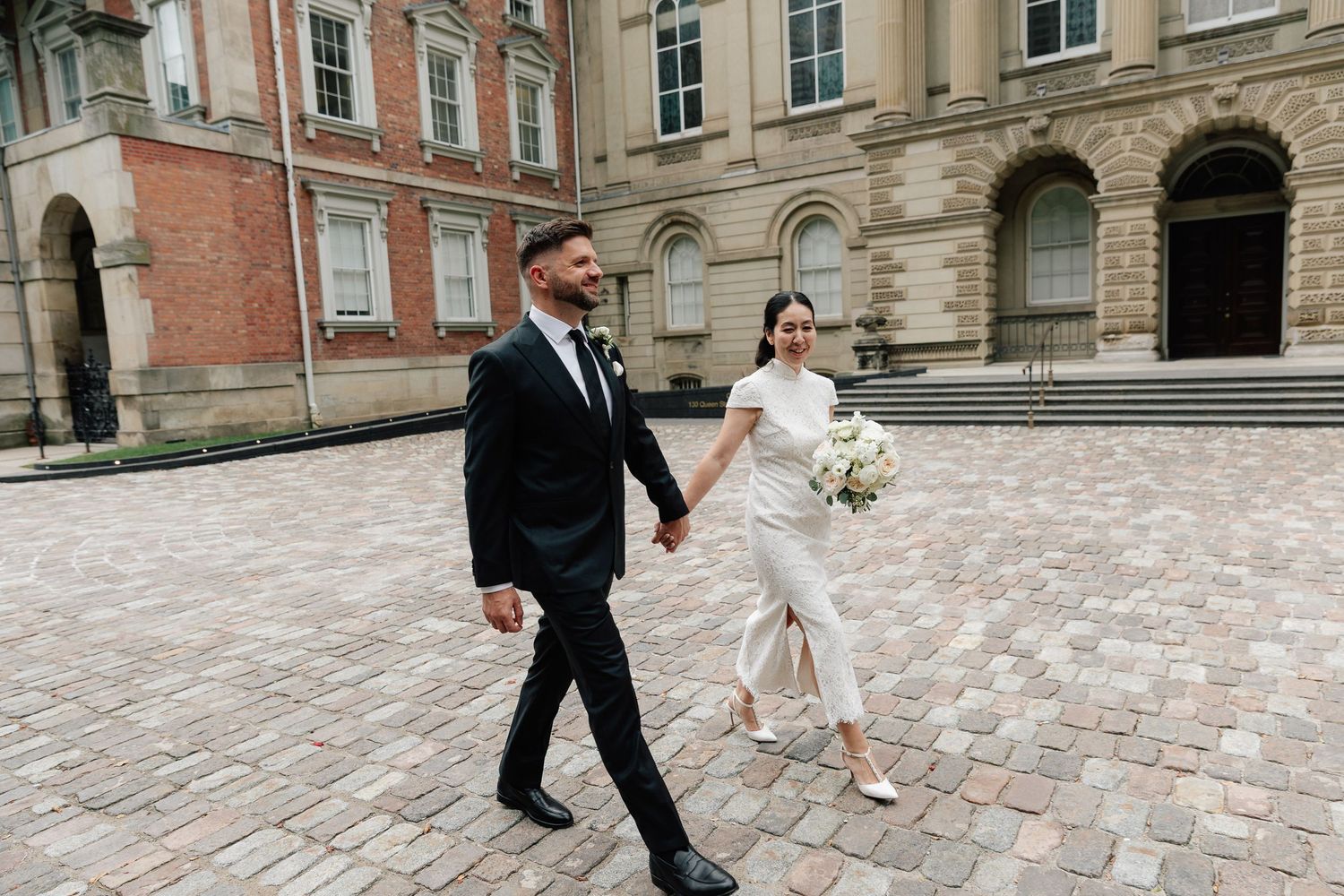 Bride in a white dress holding a bouquet walks hand in hand with a groom in a dark suit across a brick courtyard past historic buildings.