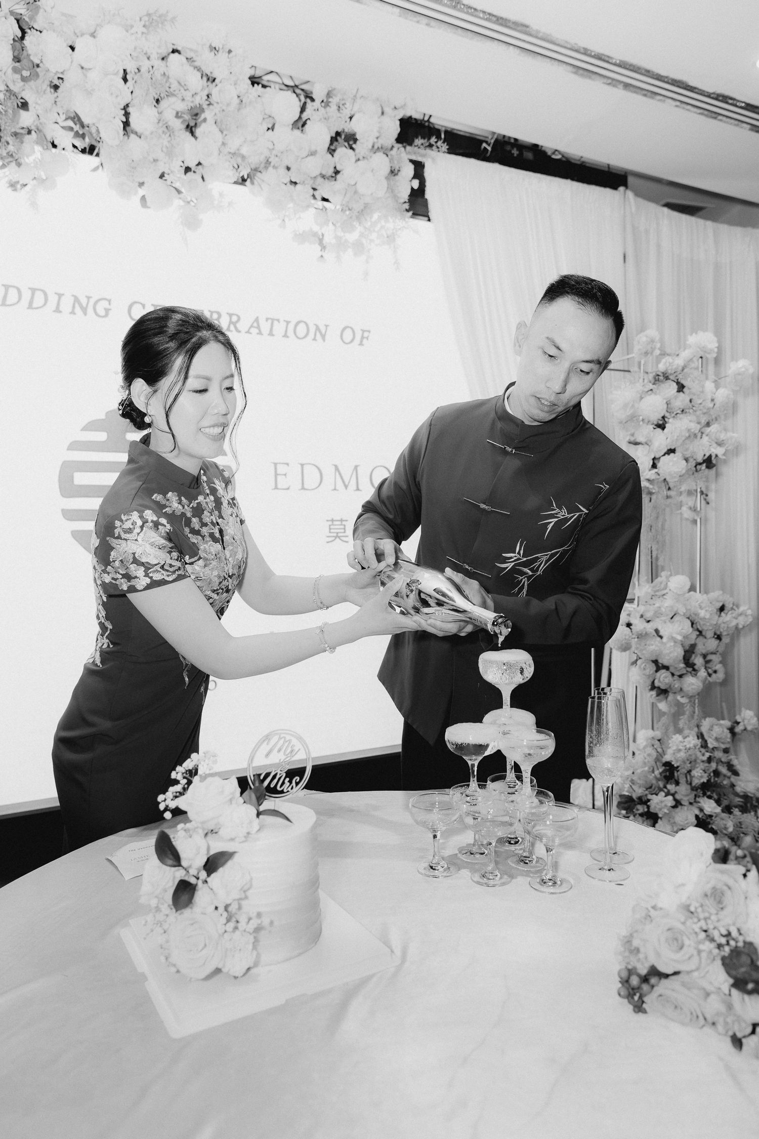 Black and white photo of a couple in traditional Chinese attire pouring champagne at their wedding celebration.