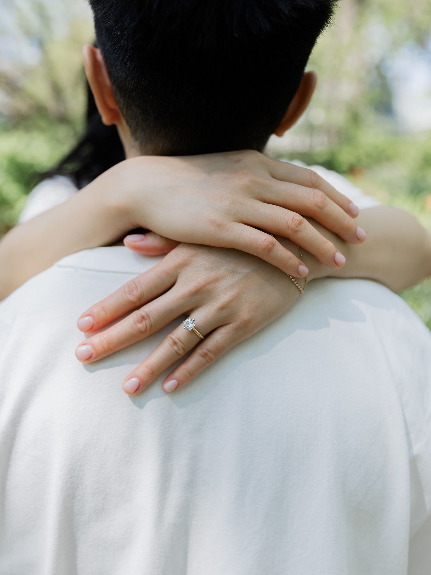 Close-up of a woman's hands with a ring wrapped around a man's shoulders in a white shirt during an outdoor engagement photoshoot.