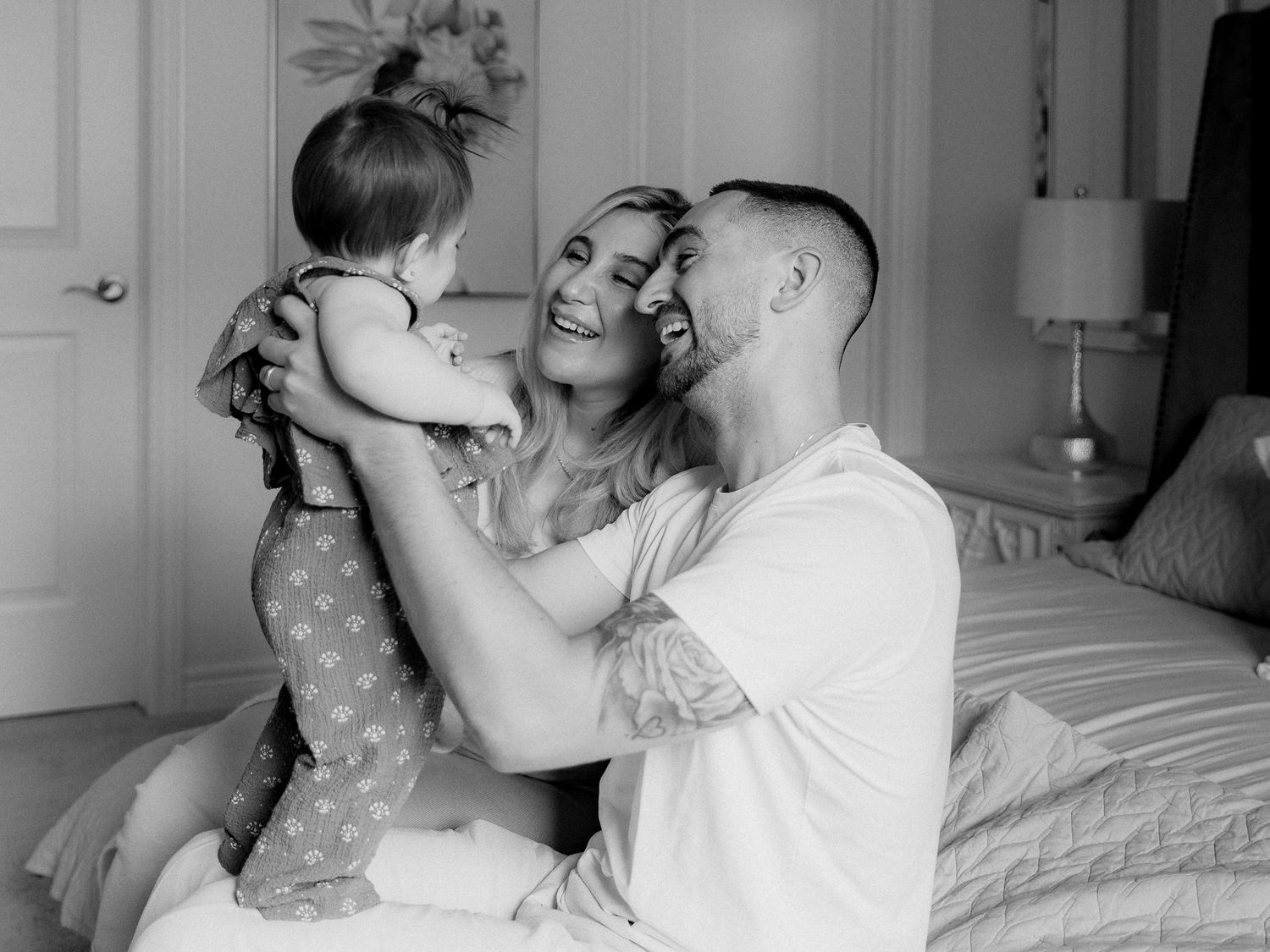 Black and white photo of a smiling couple on a bed lifting and playing with their giggling toddler in a cozy, softly lit bedroom.