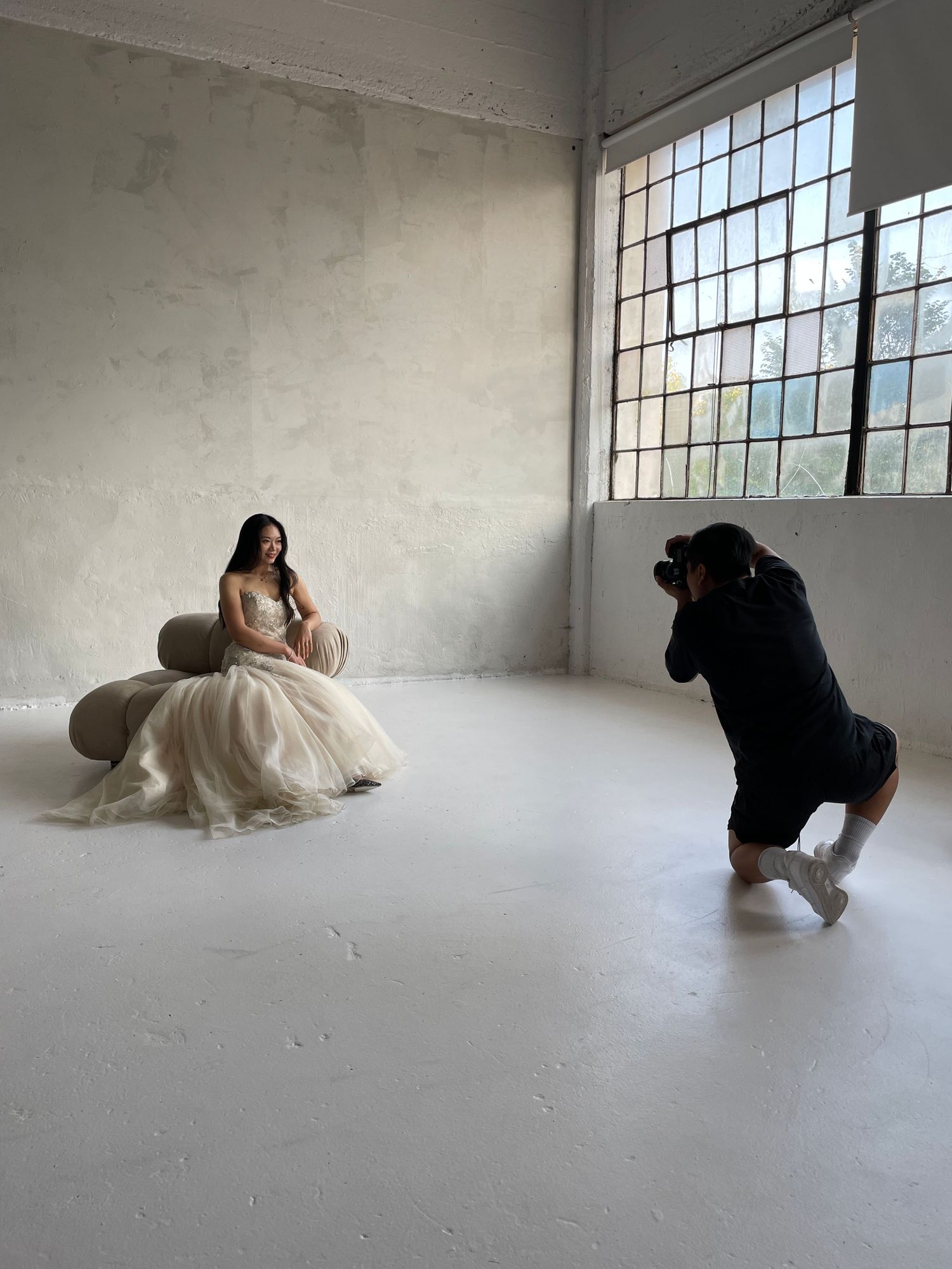 Photographer kneeling to capture a bride in a flowing gown seated in a bright studio space.