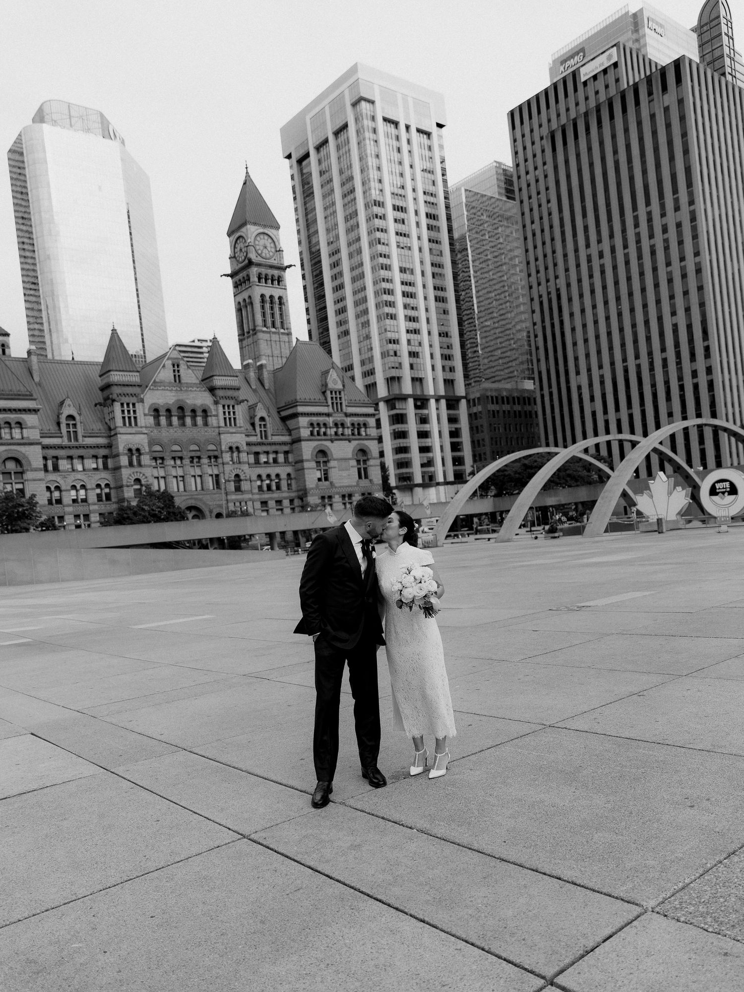 Black and white photo of a bride and groom kissing in a wide city plaza with a historic clock tower and skyscrapers behind them.