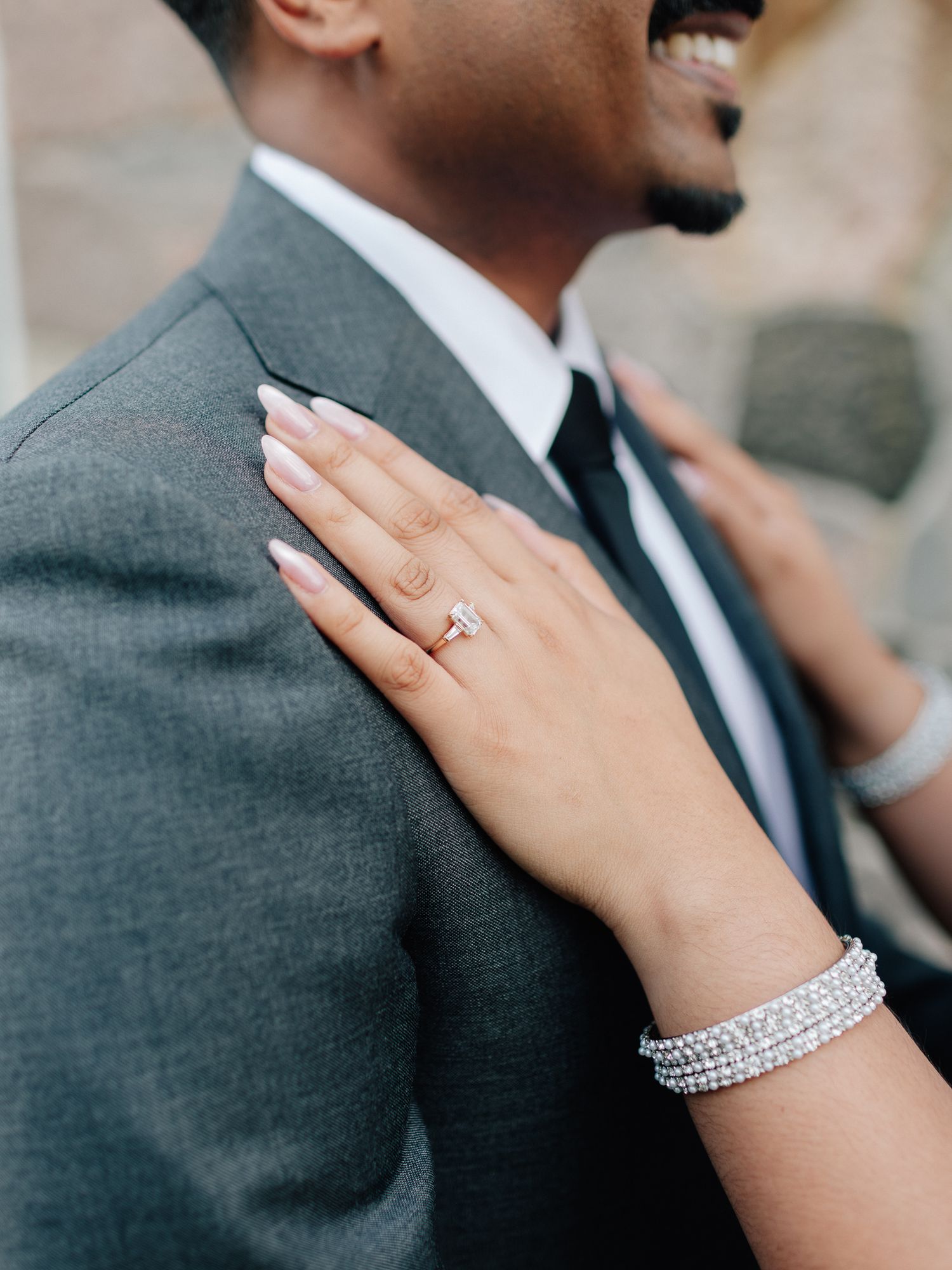 Woman's manicured hand with a sparkling engagement ring and crystal bracelet rests on a smiling man's shoulder in a gray suit.