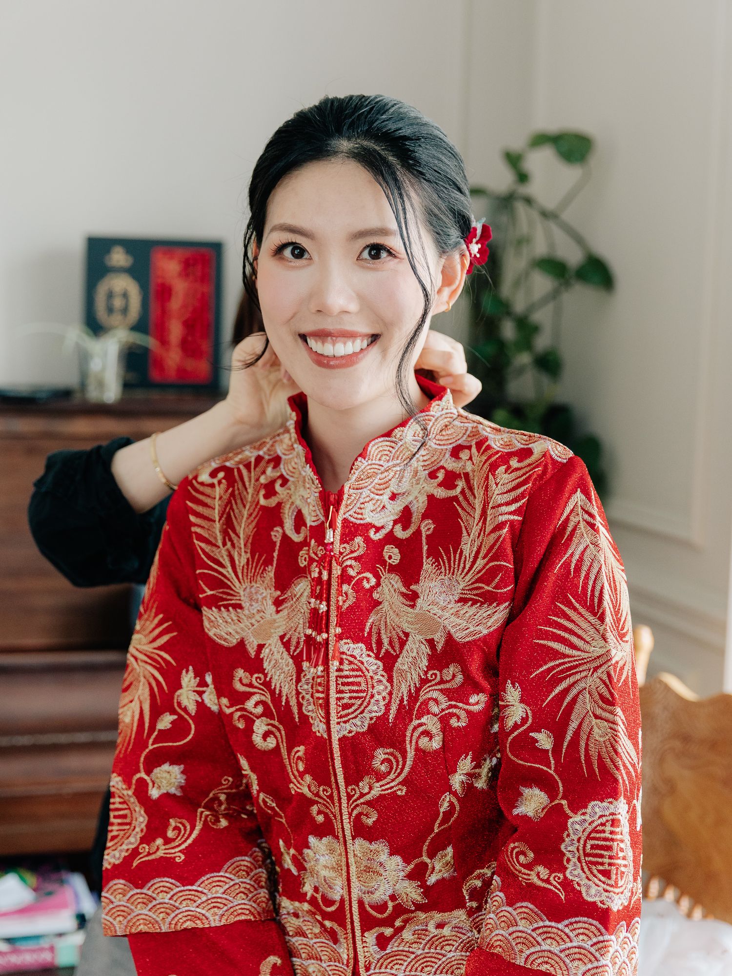 Smiling bride in a red traditional Chinese kua wedding gown with gold phoenix embroidery adjusts her hair in a bright room.