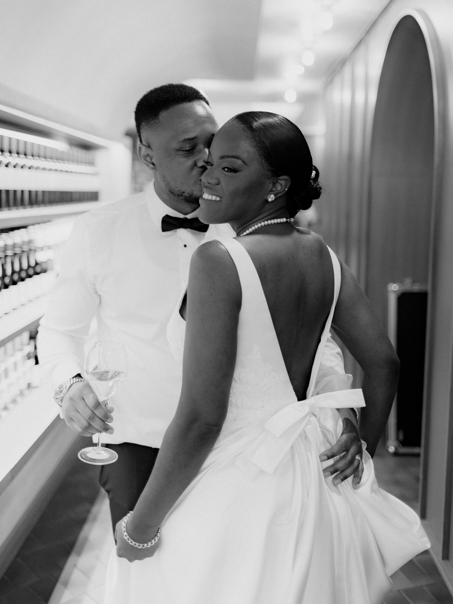 Black and white photo of a groom in a bow tie kissing his smiling bride's cheek, she wears a low-back wedding gown and pearl necklace.