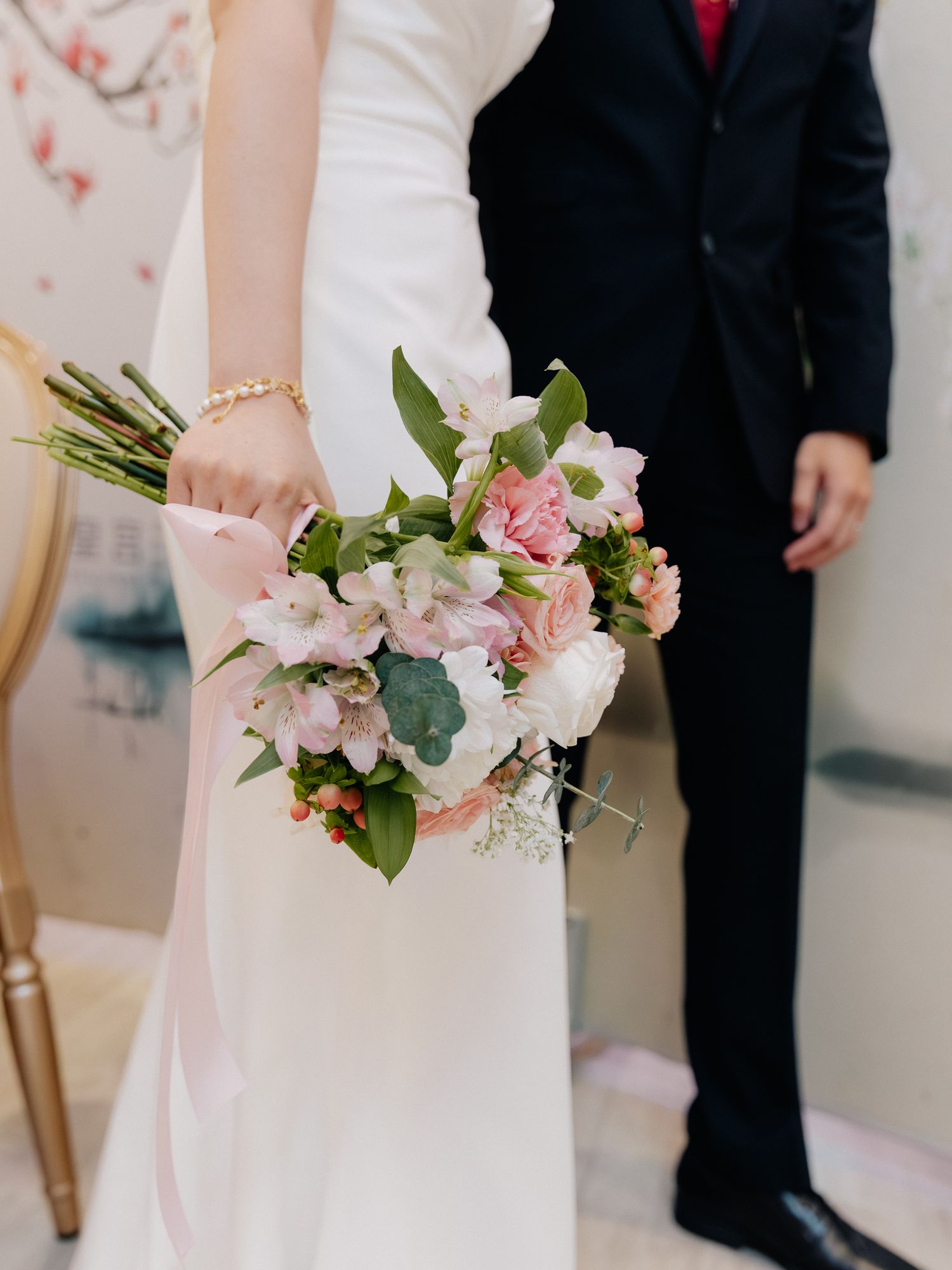 Bride in a white gown holds a pink and white floral bouquet with a ribbon beside a groom in a dark suit.