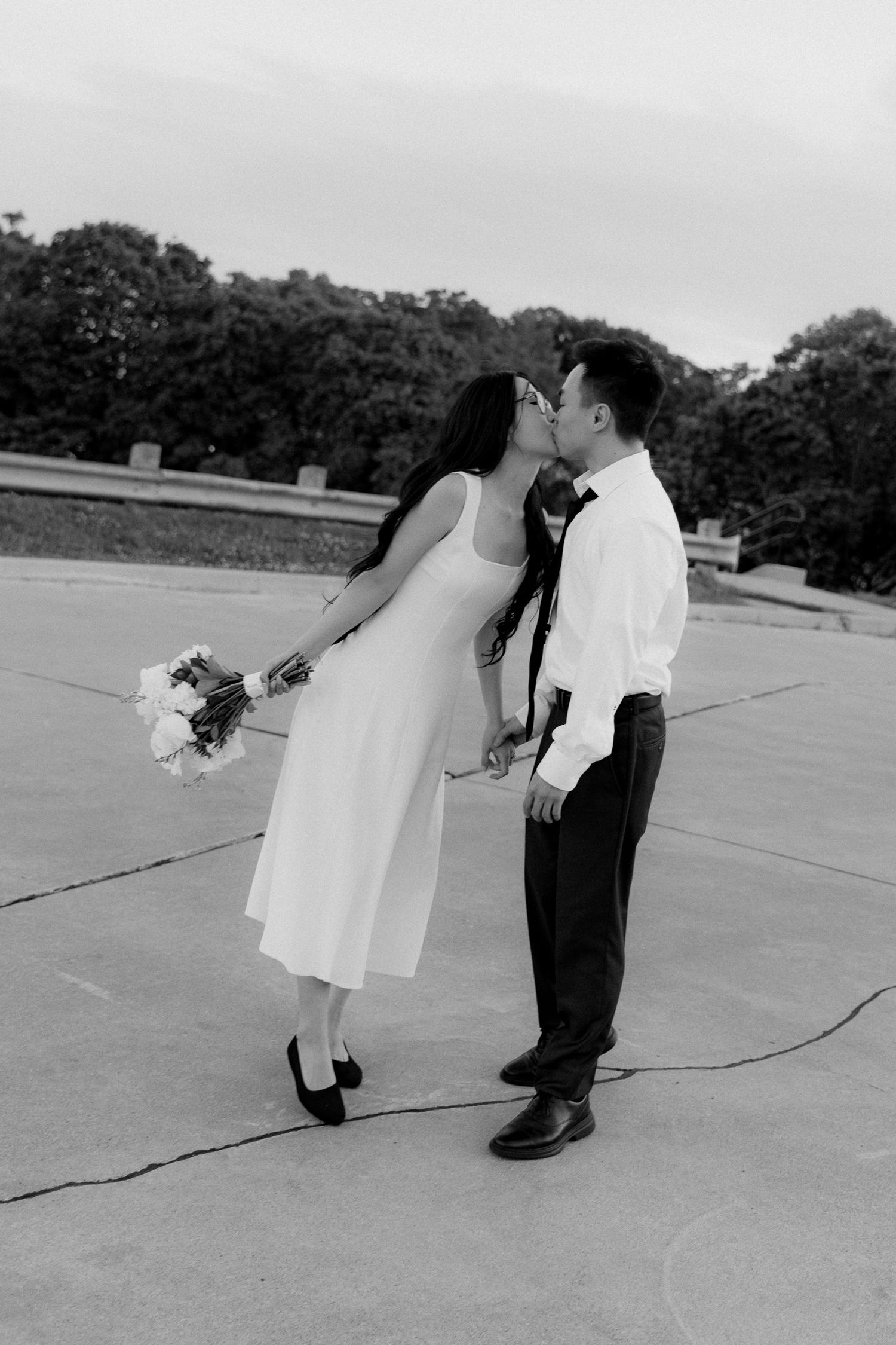 Black and white photo of a couple kissing on a cracked pavement road, the woman in a white midi dress holding a bouquet of flowers.