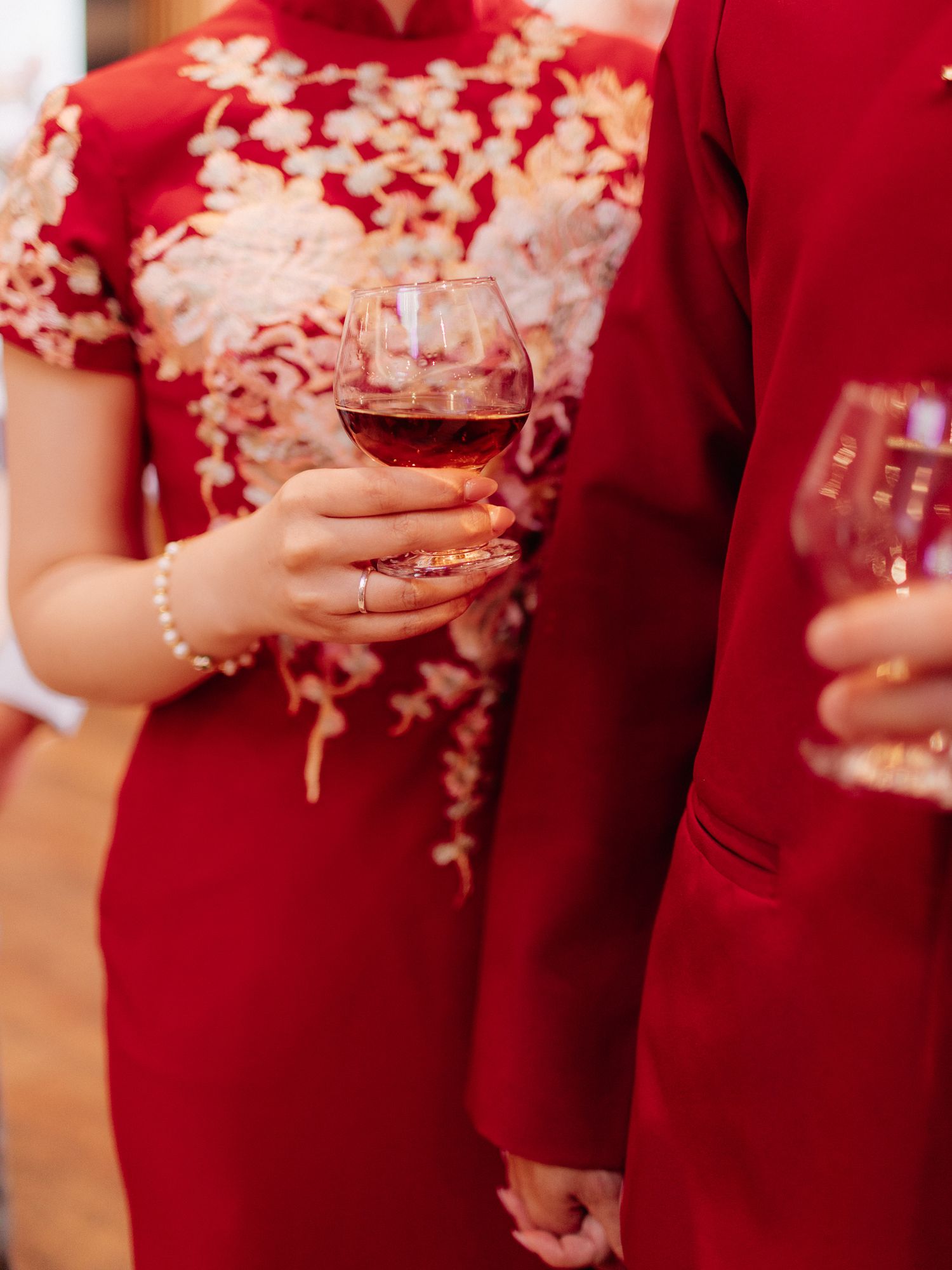 Woman in a red qipao with gold floral embroidery holds a glass of wine beside a person in a red jacket at a celebration.