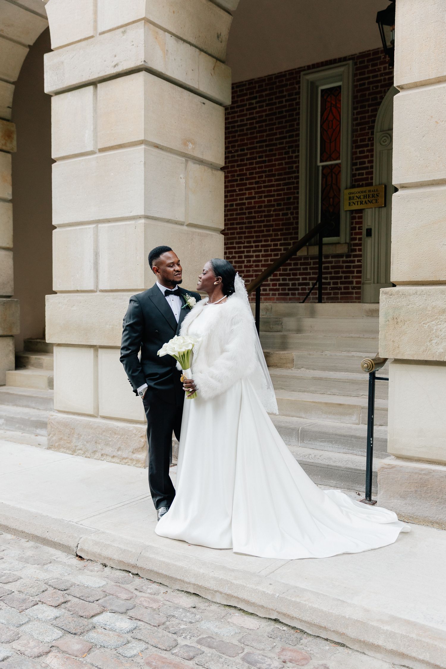 Bride in a white gown with a fur stole and groom in a black tuxedo hold a bouquet on stone steps outside a brick and limestone building.