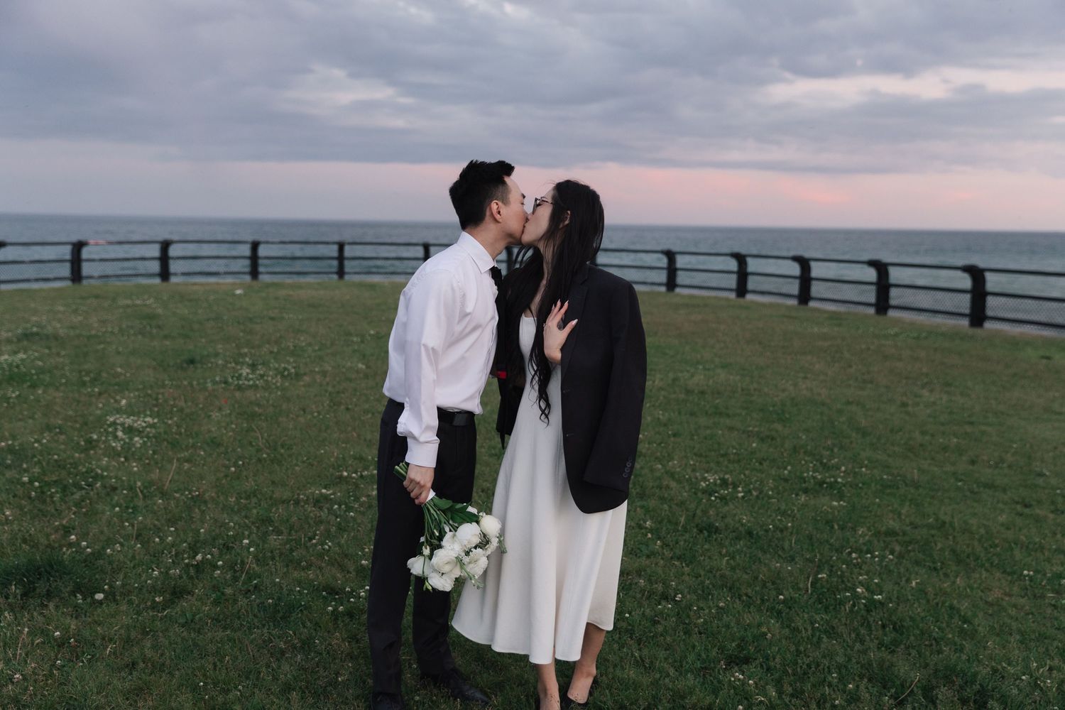 Couple kissing on a grassy seaside lawn at dusk, the bride in a white dress and black blazer holding a white flower bouquet.