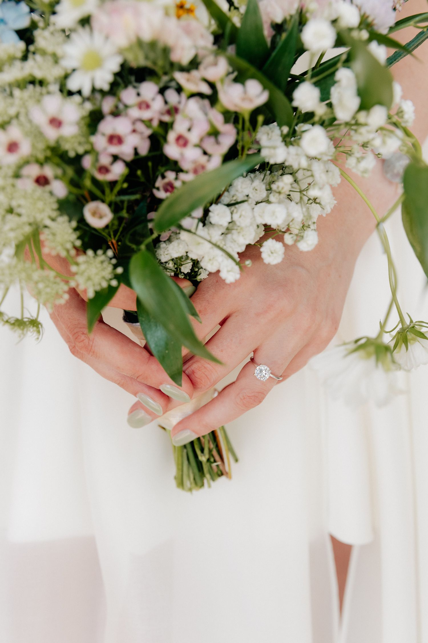 Bride in a white dress holds a wildflower bouquet of baby's breath and pink blooms, showing a diamond halo engagement ring.