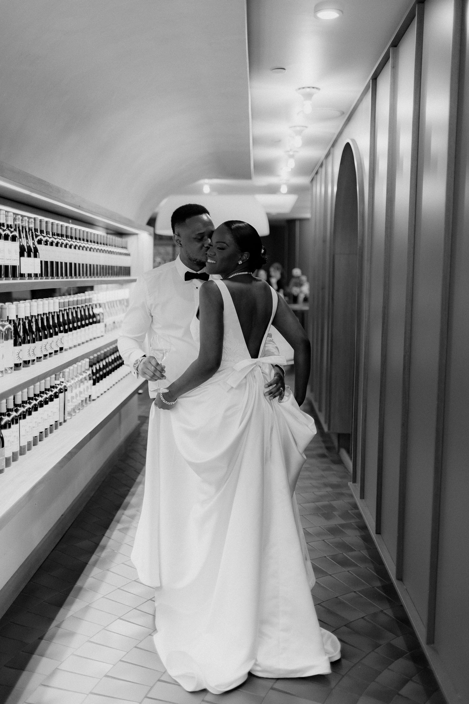 Black and white photo of a bride in an open-back gown embracing her groom in a bow tie in an arched hallway lined with wine bottles.