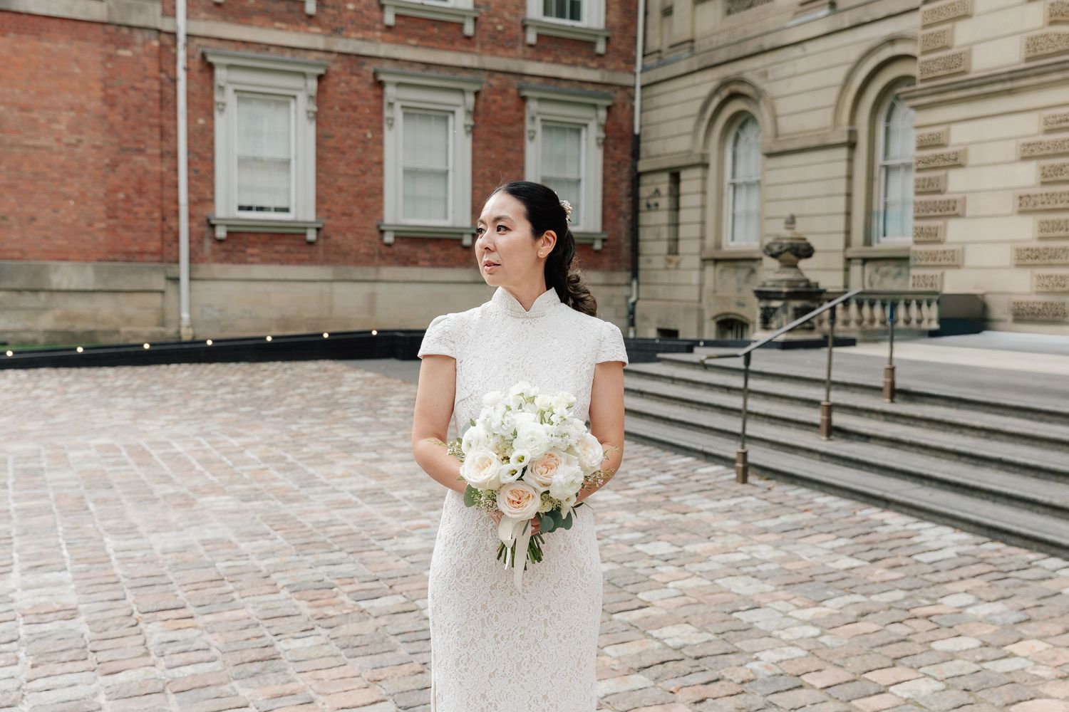 Bride in an elegant white lace dress holds a bouquet of white and blush flowers on a brick courtyard outside a historic building.