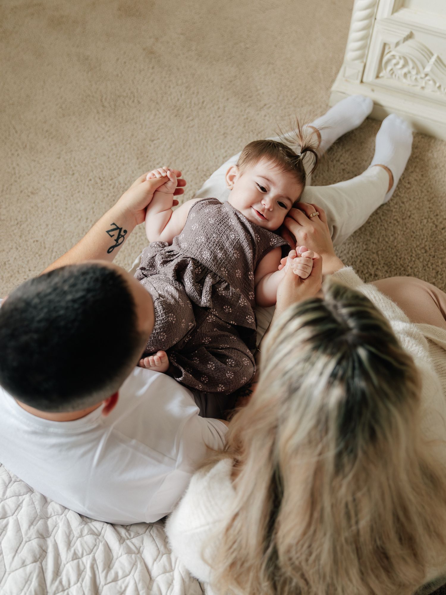 Smiling baby in a brown romper lies on a quilted blanket, holding hands with two parents seen from above in a warm family moment.
