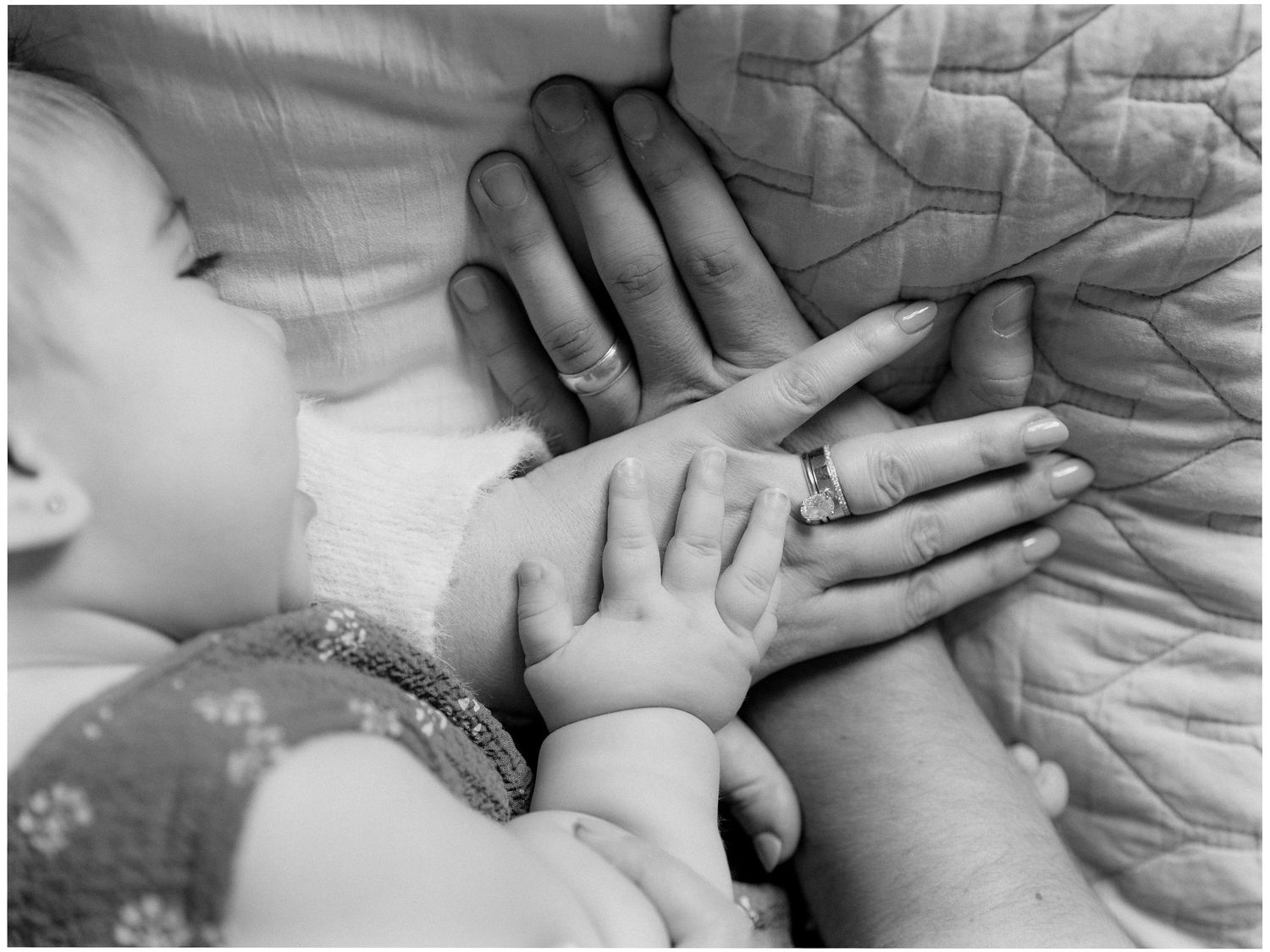 Black and white photo of a baby's tiny hand resting on an adult's hands atop a quilted blanket, capturing a tender parent and child moment.