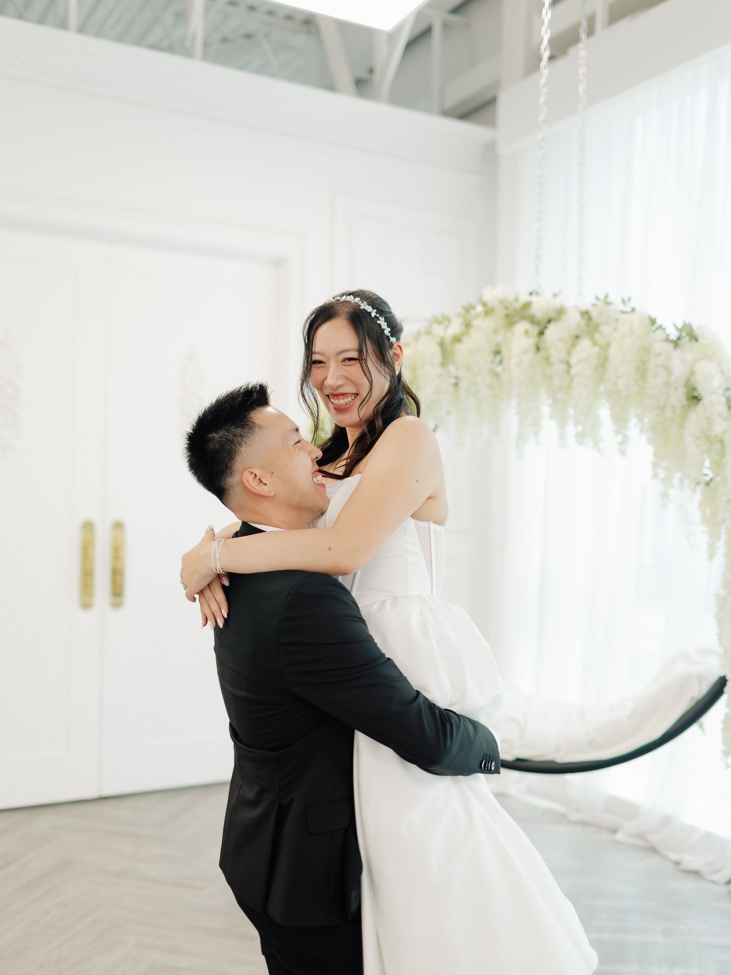 A groom in a black suit lifts his smiling bride in a white wedding gown beside a white floral arch in a bright, elegant venue.