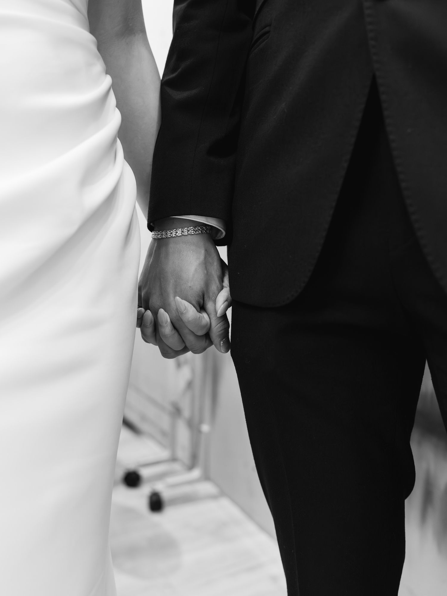 Black and white photo of a bride and groom holding hands, showcasing a diamond bracelet and wedding attire.