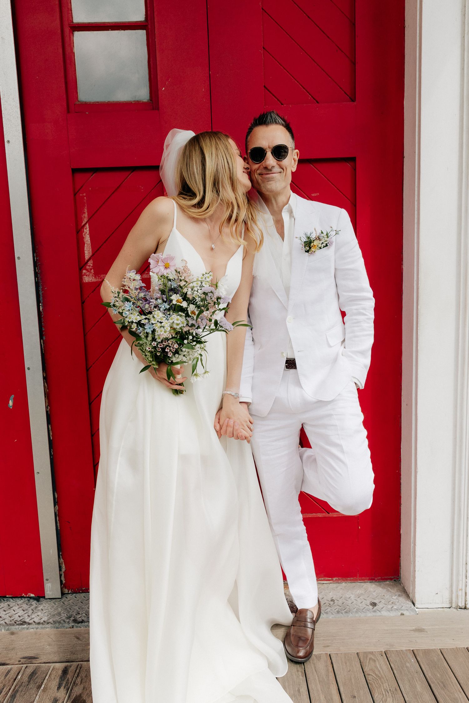 Bride in a white gown holding a wildflower bouquet poses hand in hand with a groom in a white suit and sunglasses by a red barn door.