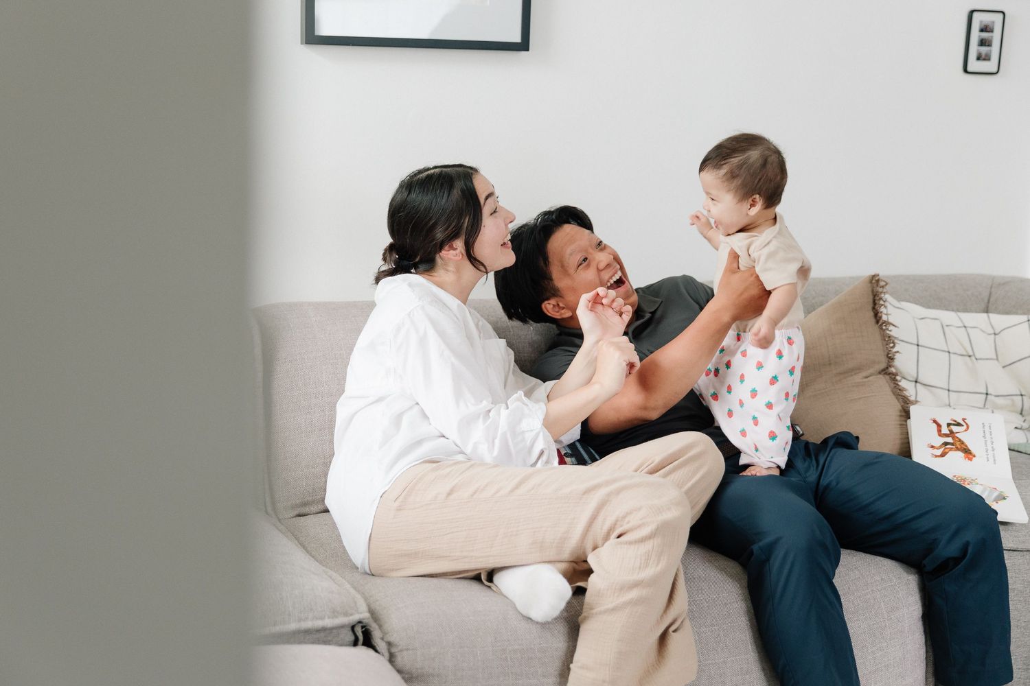 Smiling parents sit on a gray sofa in a bright living room, playfully lifting their giggling baby between them.