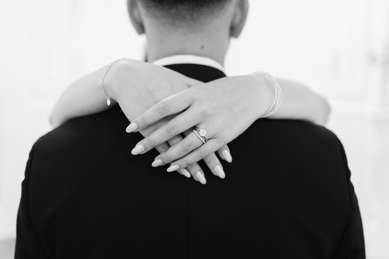 Black and white photo of a bride's hands with a diamond engagement ring wrapped around the shoulders of a groom in a dark suit.