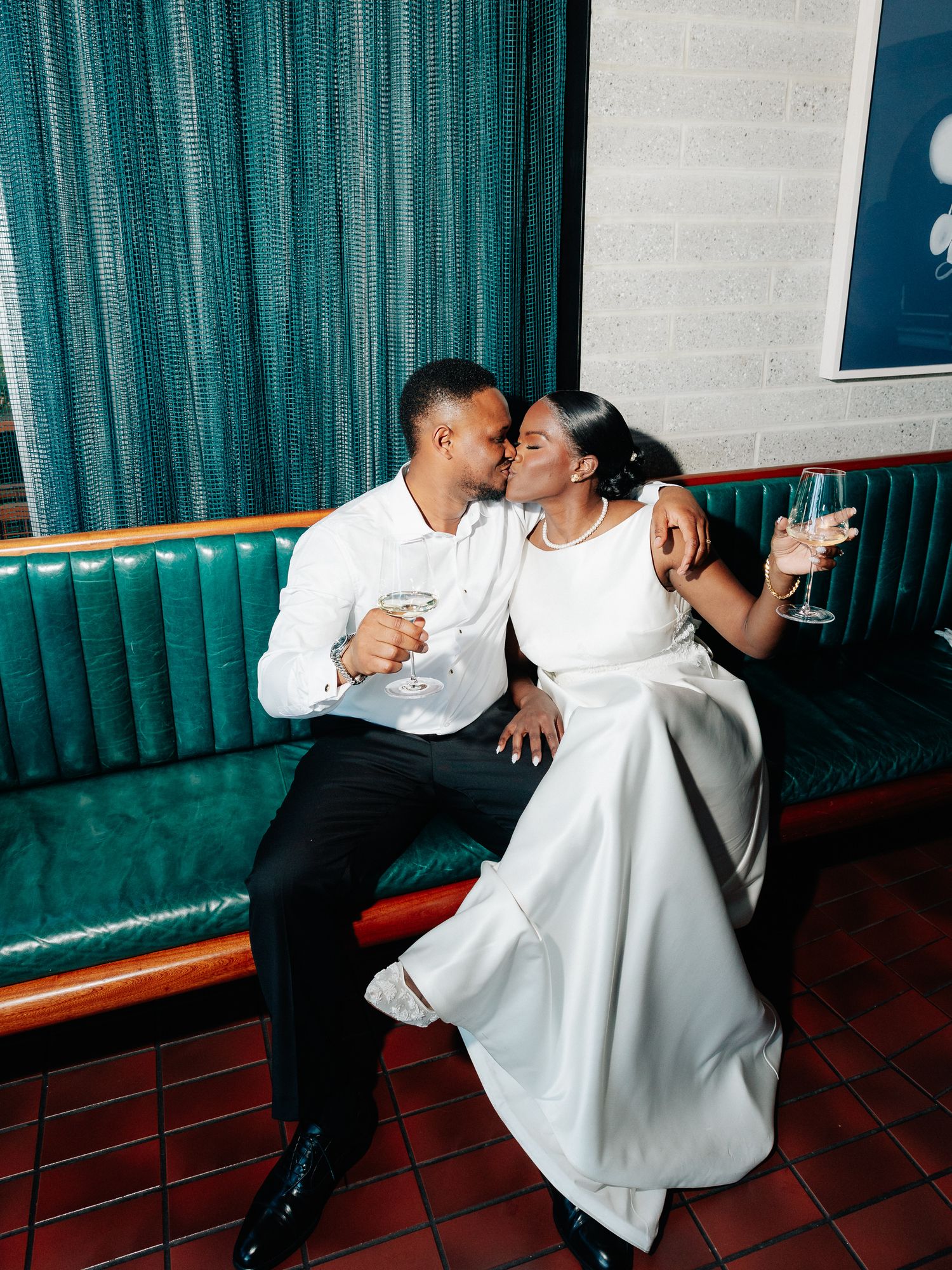Bride in white gown and groom in white shirt kiss on a green tufted leather booth while holding champagne glasses.
