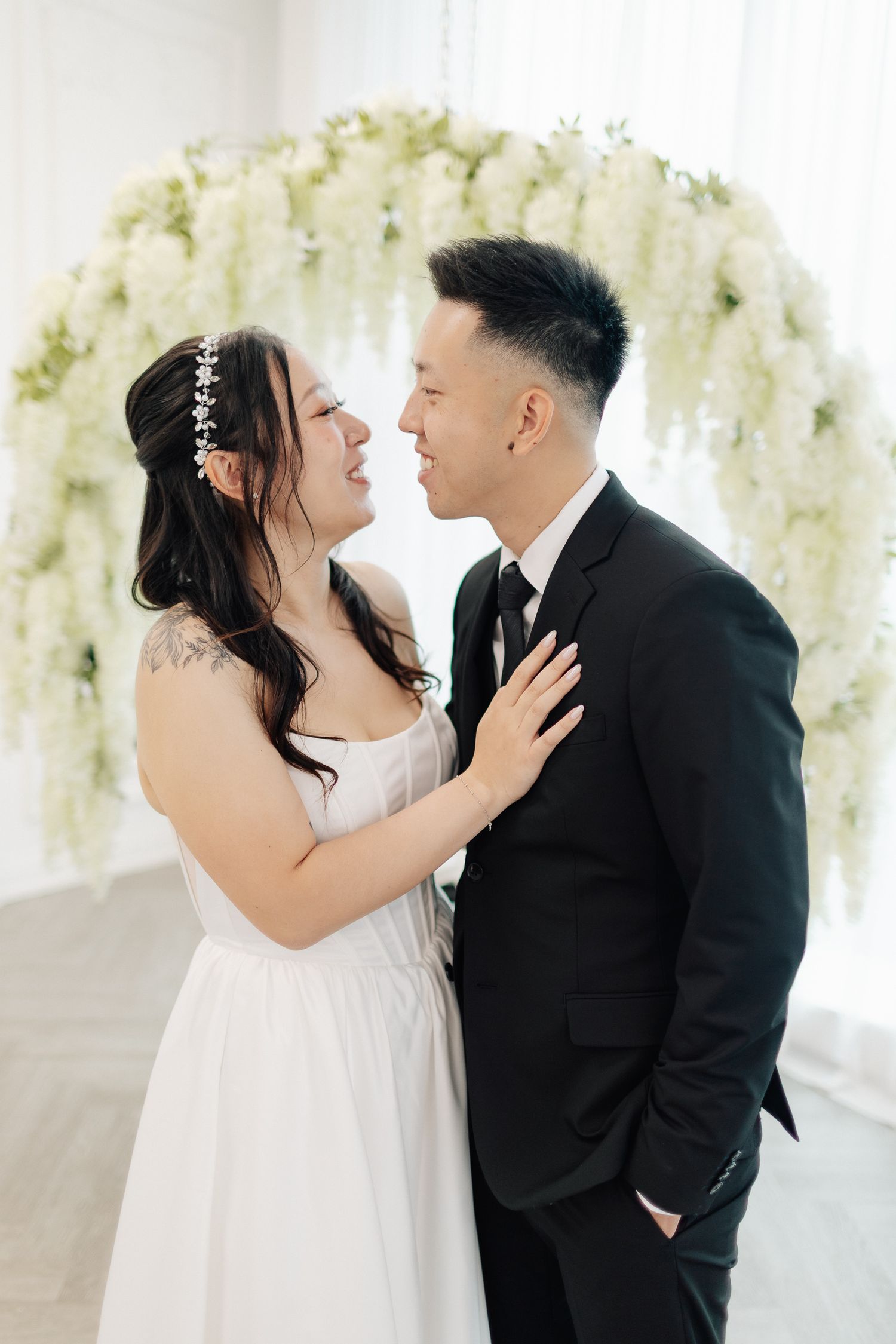 Smiling bride in a white gown and floral headpiece embraces a groom in a black suit beneath a white wisteria flower arch.