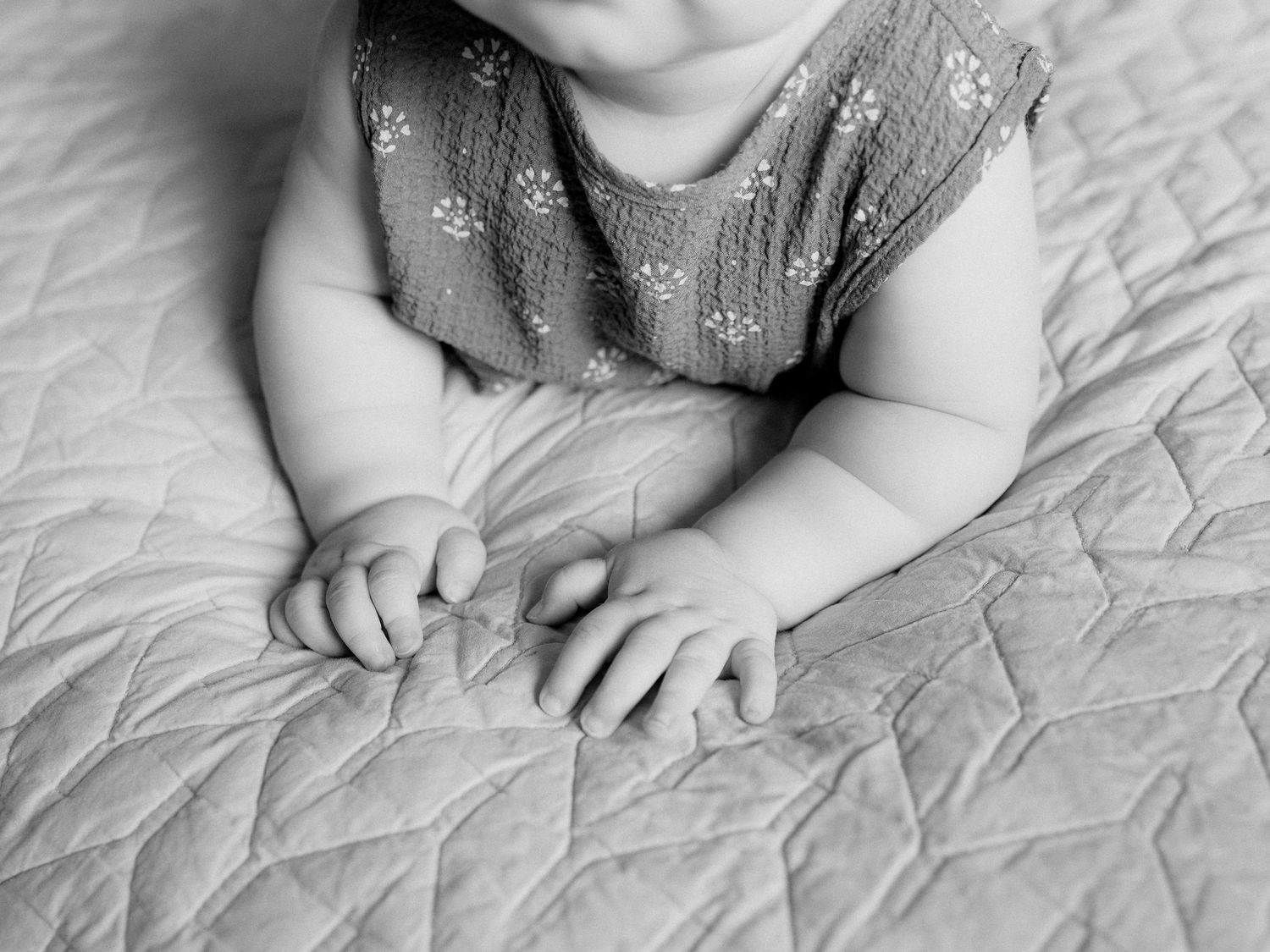 Black and white photo of a baby in a floral sleeveless top lying on their tummy on a quilted blanket with hands resting on the fabric.