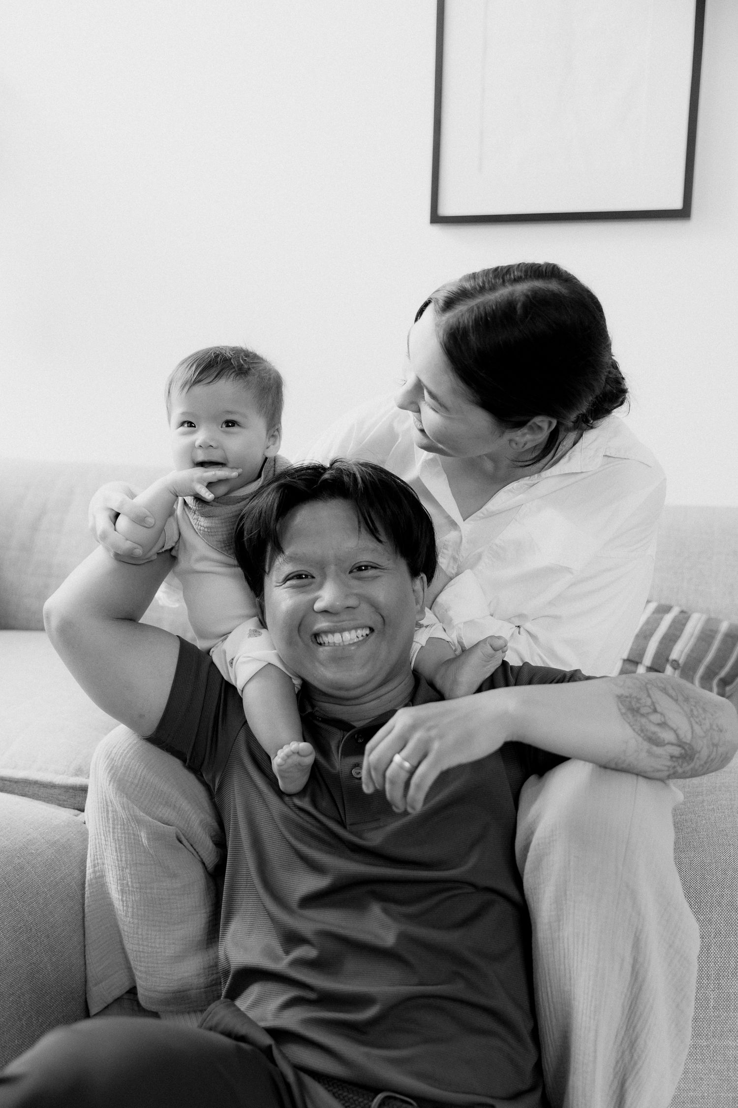 Black and white photo of a smiling man on a sofa with a baby on his shoulders and a woman beside him, enjoying a cozy family moment.