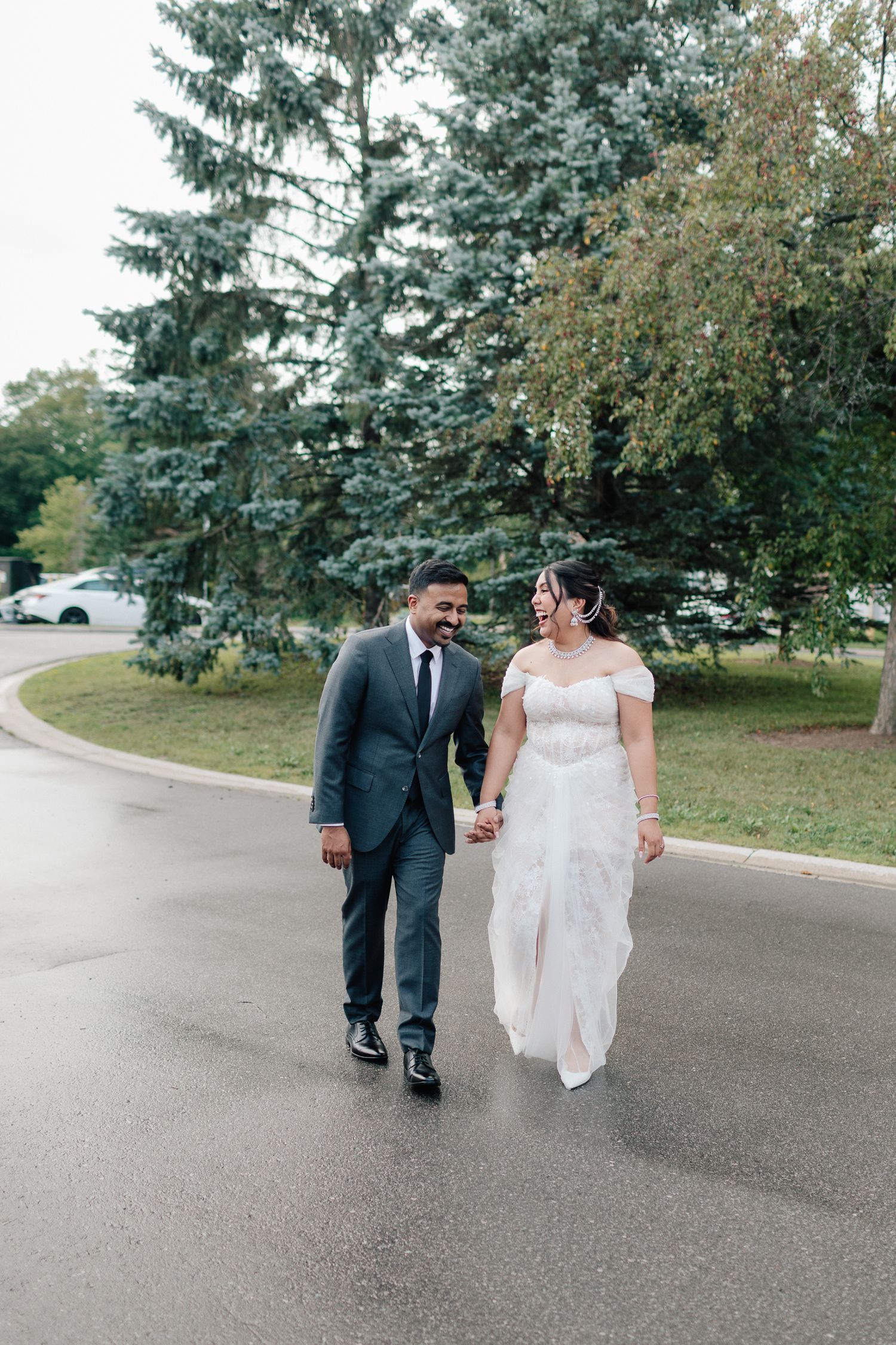 Bride in a white off-shoulder gown and groom in a gray suit hold hands and smile while walking down a wet road lined with evergreen trees.