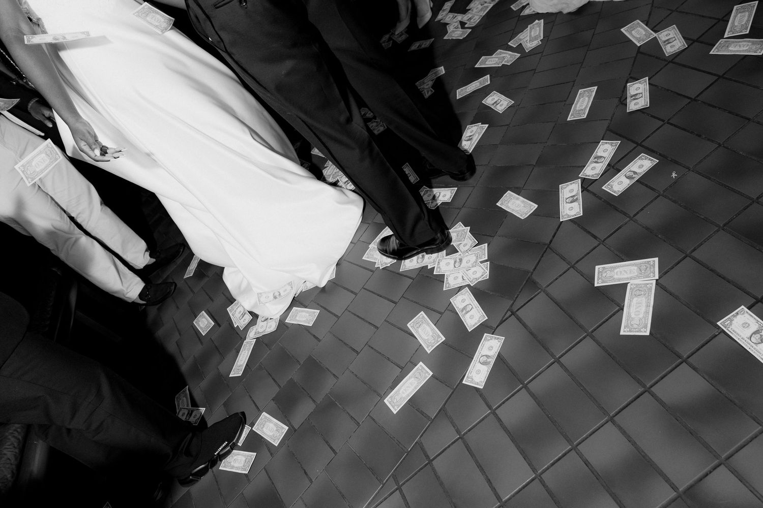 Black and white photo of a bride in a white gown and guests standing on a tiled floor covered in scattered paper money.