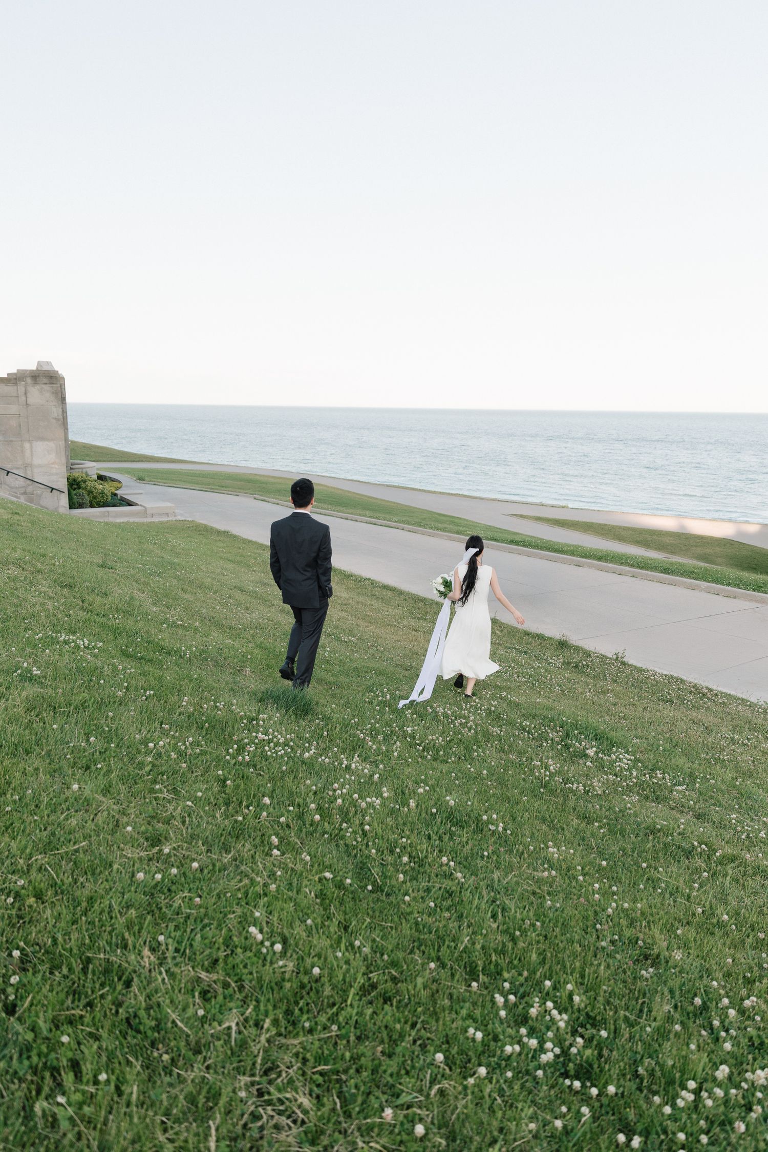 Bride in a white gown and groom in a dark suit walk across a grassy hillside toward a paved path overlooking a calm lakeshore.